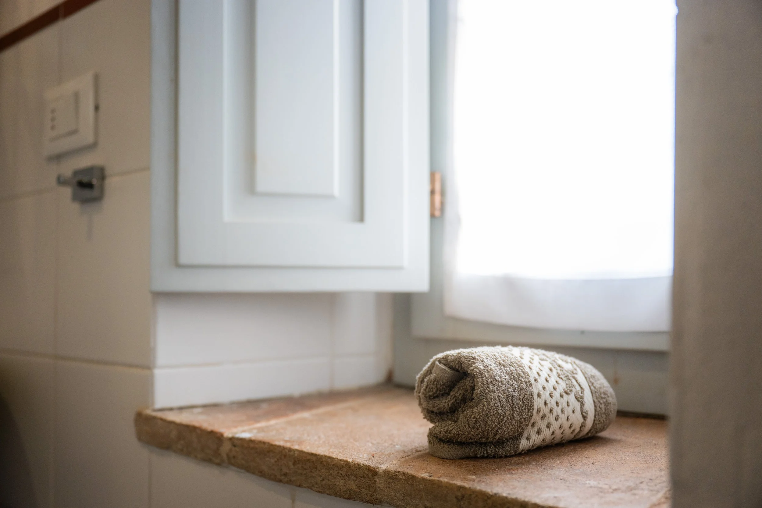 Asciugamano arrotolato sulla finestra con tende bianche, pareti di ceramica bianca e davanzale in pietra.