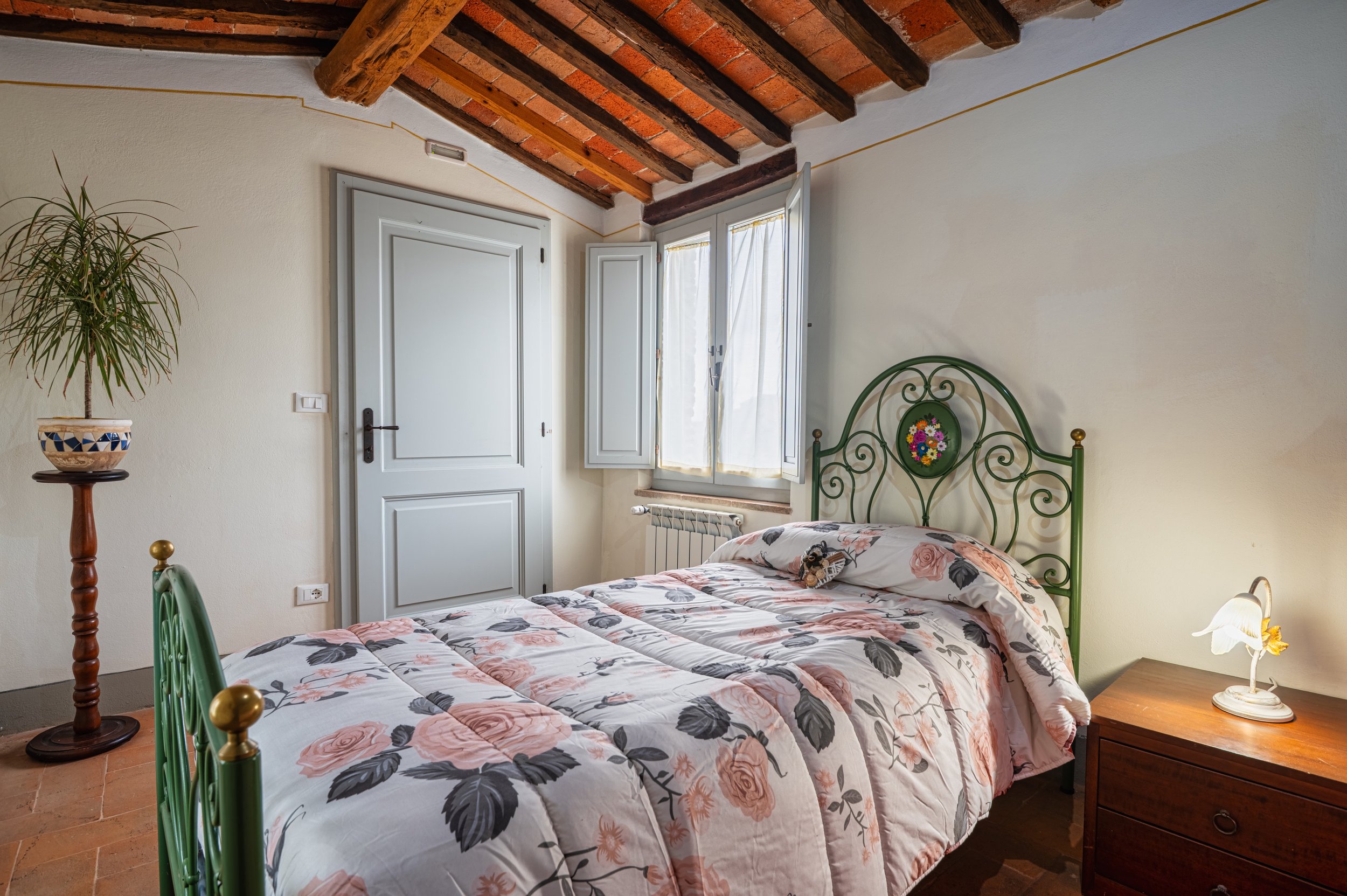 Camera da letto con letto a baldacchino, coperto con una trapunta floreale rosa e grigia, lampada accesa sul comodino di legno, finestra con tende leggere, pareti chiare, soffitto con travi in legno.