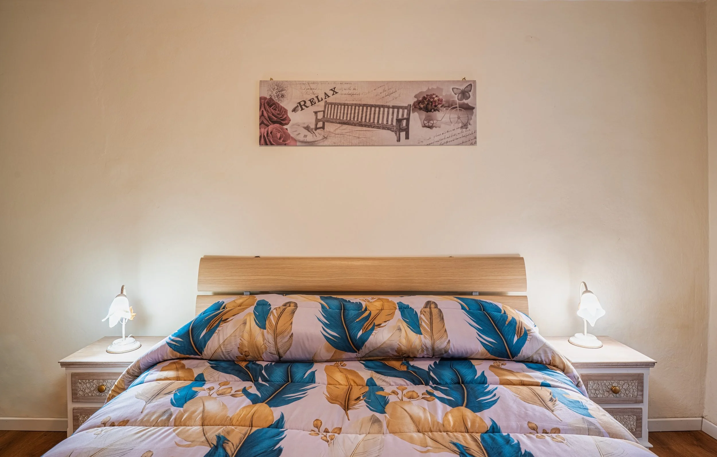 Letto con testiera di legno naturale, lenzuola e cuscini con motivo a foglie blu e beige, due lampade ai lati, quadro in bianco e rosa con panchina, rose e farfalle.