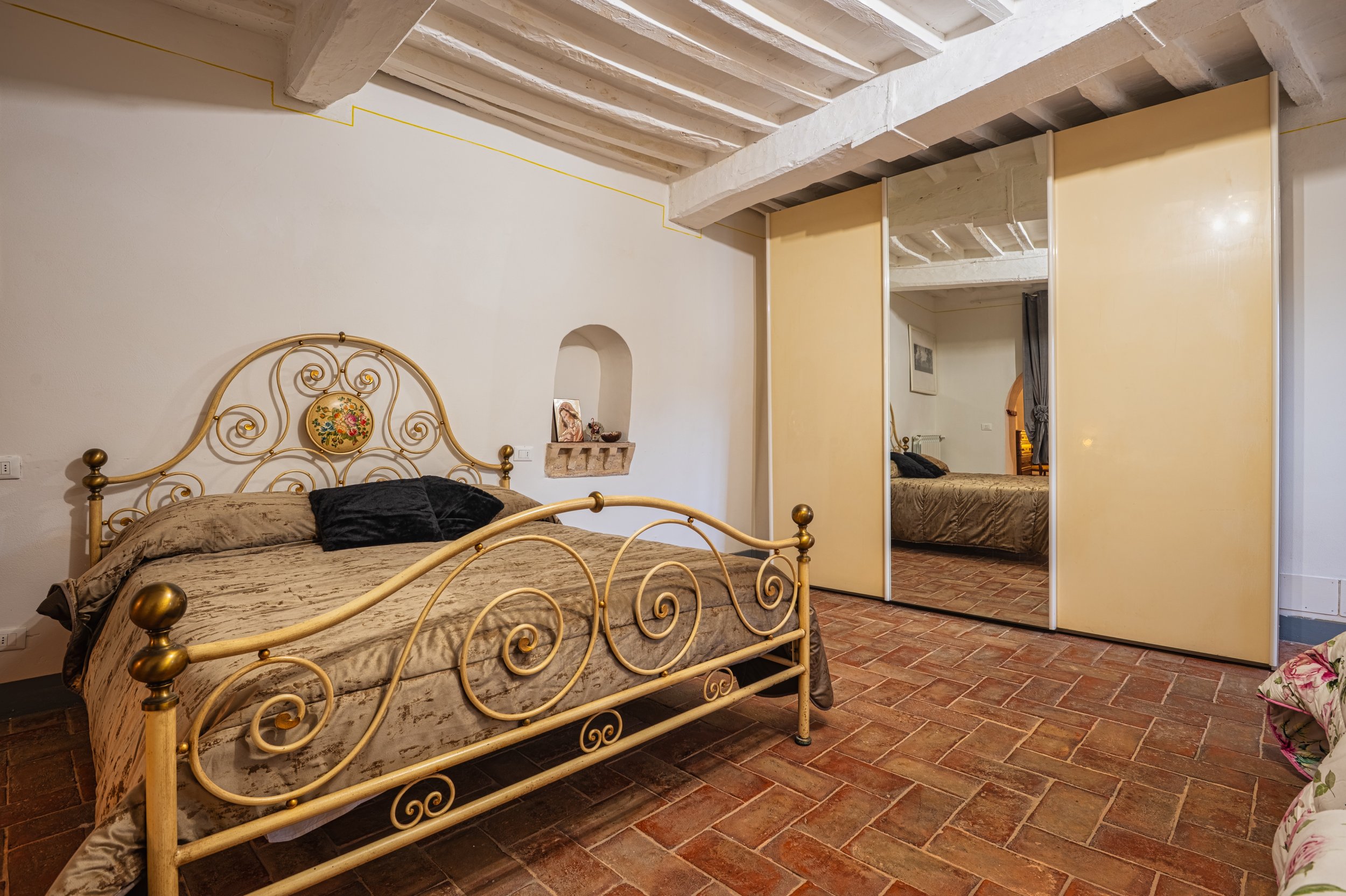 Camera da letto con letto in ferro battuto dorato, letto con copriletto beige e cuscini neri, armadio con specchi a porte scorrevoli, muro bianco, pavimento di mattonelle di terracotta, soffitto con travi di legno.