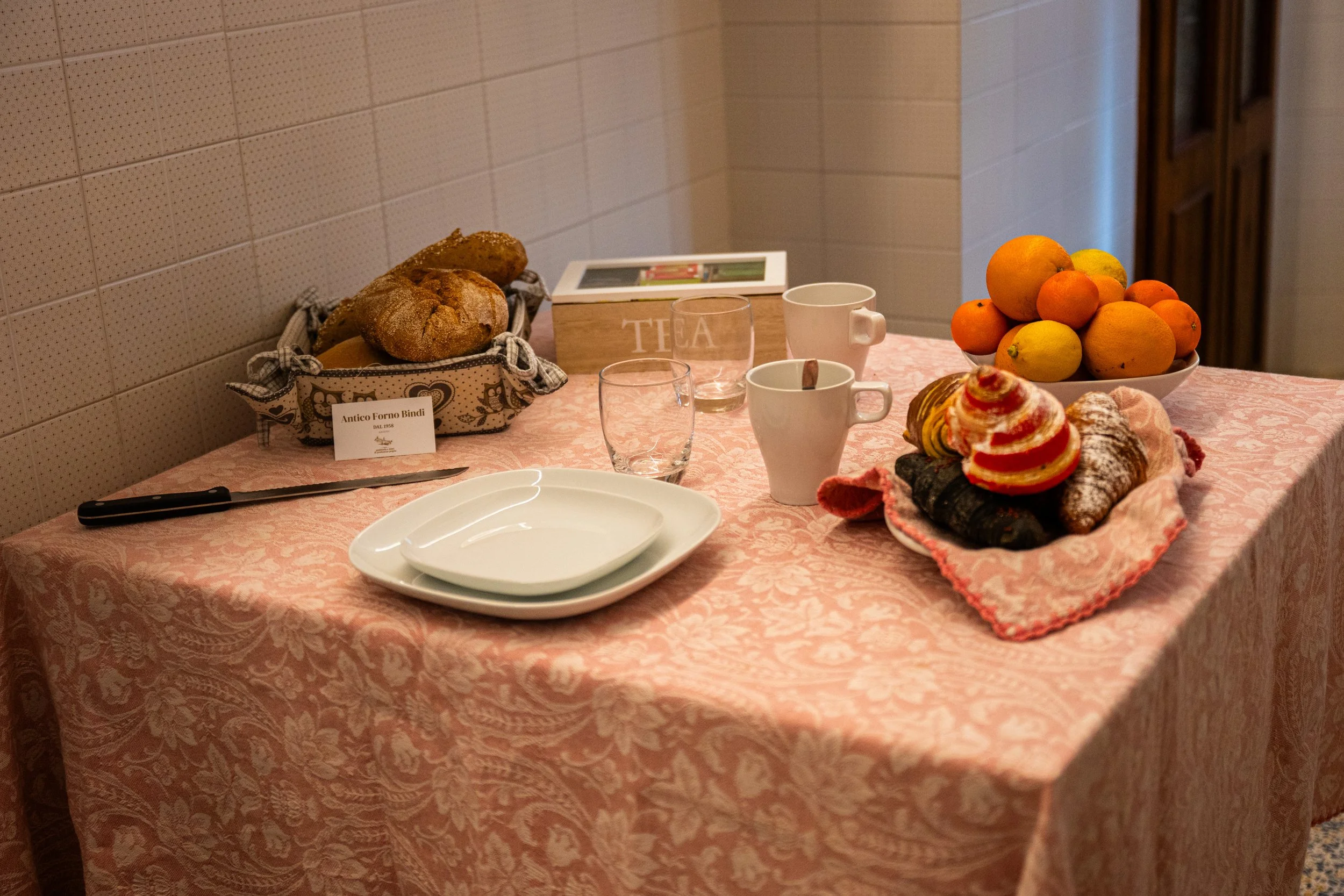 Tavolo apparecchiato con pane, tazze, bicchieri, una ciotola di agrumi, una ciotola di dolci e piatti. Sul lato sinistro, un coltello e un segnalino con scritto "Antico Forno Bindi". Sullo sfondo, una scatola con la parola "TEA".