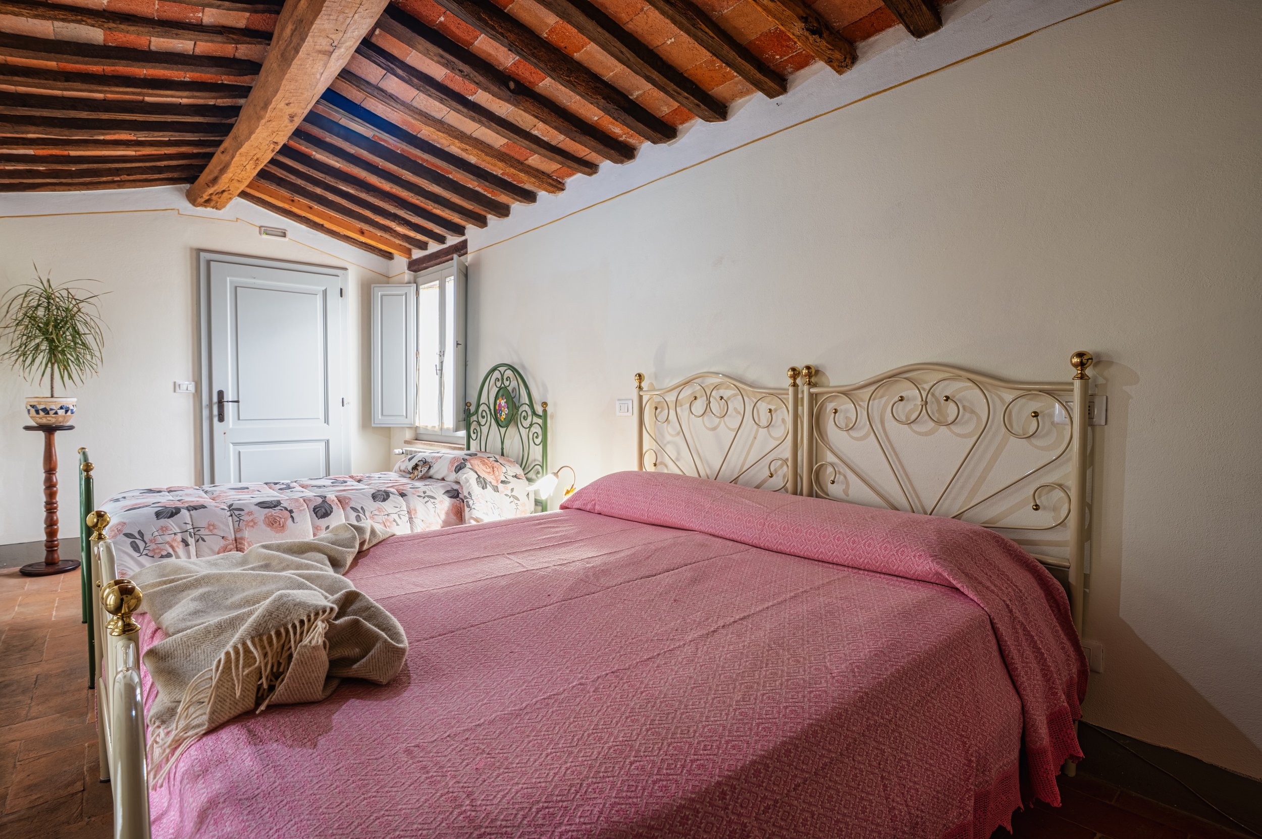 Camera con soffitto a travi di legno e due letti, uno matrimoniale con testa in ferro battuto decorata, e uno singolo con copriletto floreale, finestra aperta con luce naturale