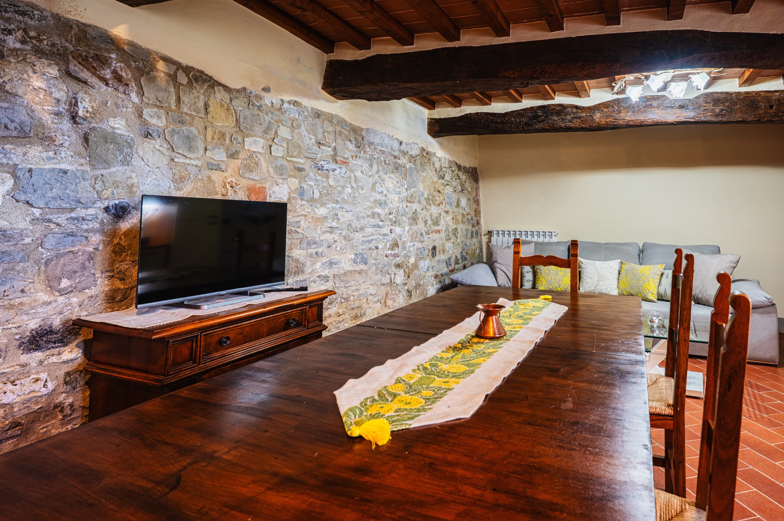 Interno di una stanza con tavolo di legno, divano grigio con cuscini, parete in pietra, soffitto con travi di legno, televisore su mobile di legno, e lampadario con luci in alto.