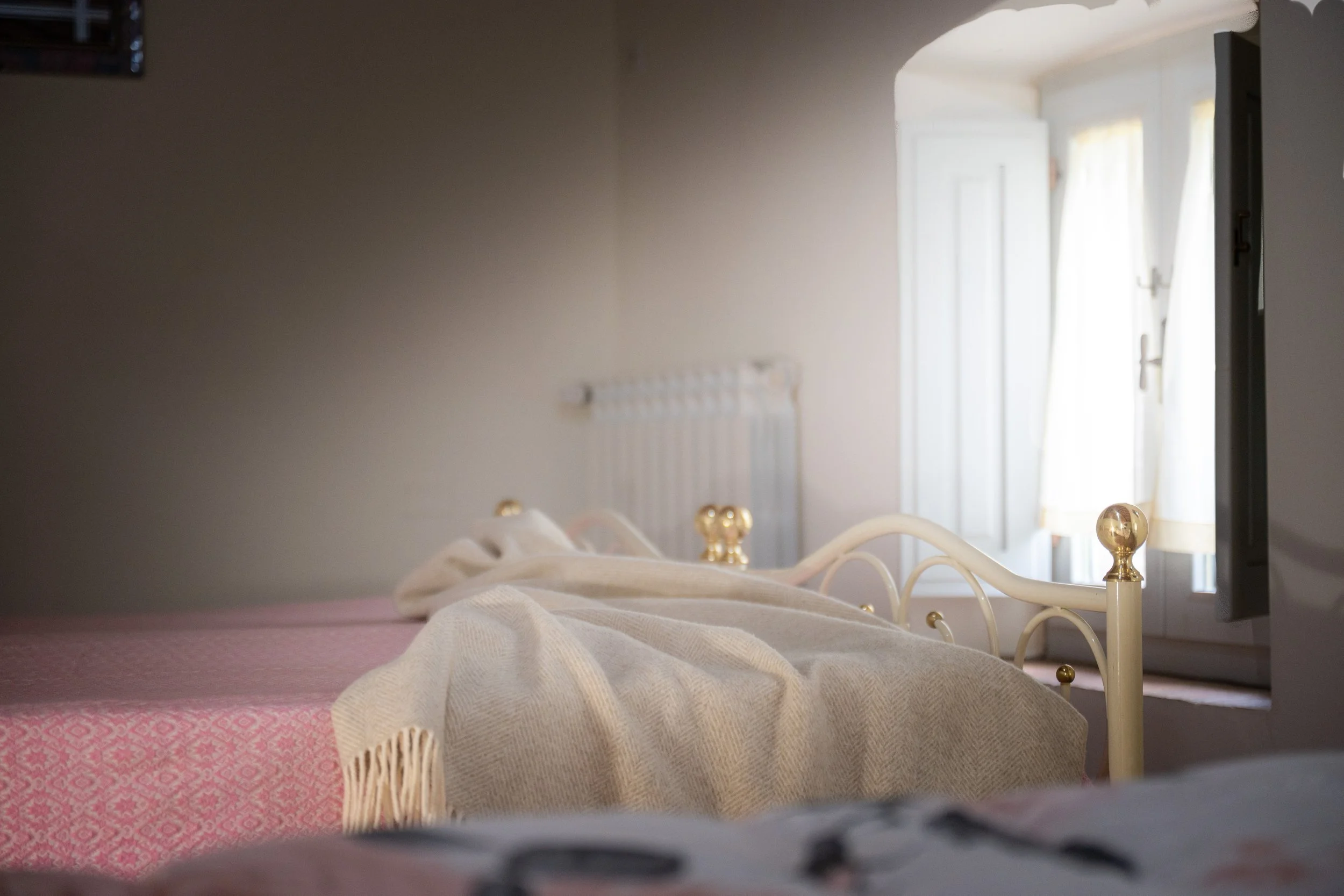 Letto con coperta beige in una stanza con finestra bianca e tendaggi leggeri.