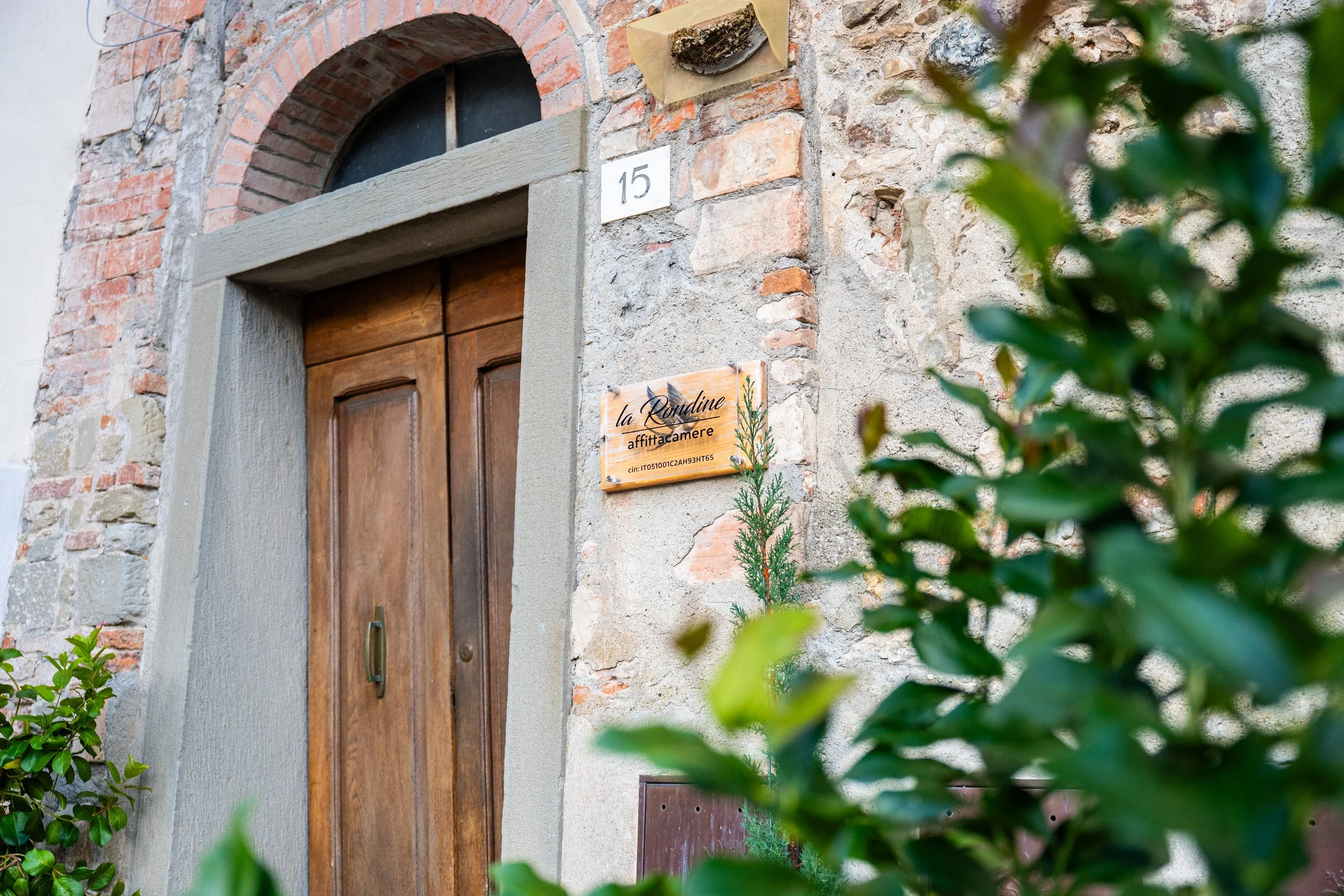 Porta in legno con cornice di pietra e muro di pietra e mattoni, con un cartello che dice 'la Rondine affittacamere' e il numero 15, circondato da piante verdi.