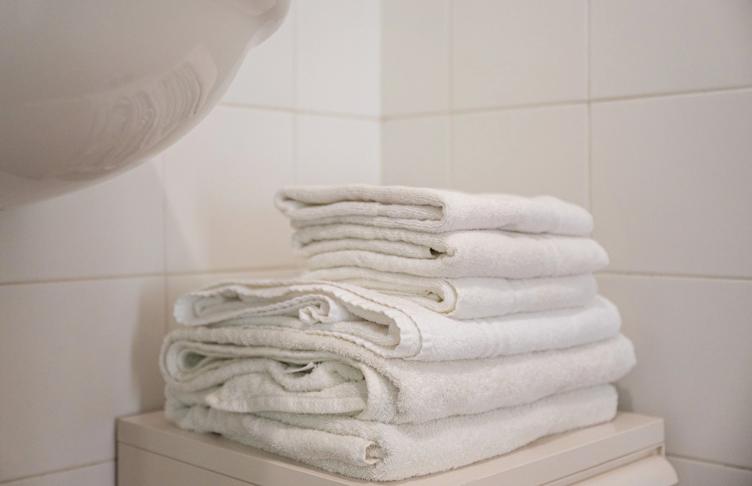 Asciugamani bianchi piegati uno sopra l'altro su una superficie in un bagno con pareti rivestite di piastrelle bianche.