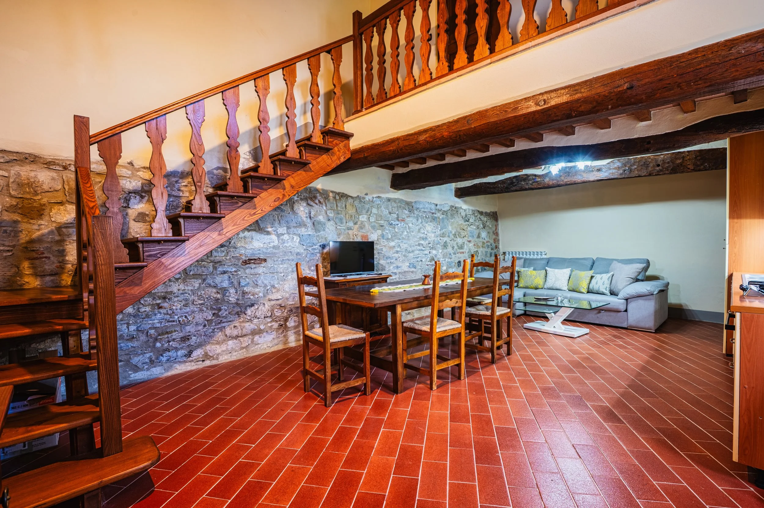 Interno soggiorno con parete in pietra, scala in legno, tavolo in legno con sedie, divano grigio, tavolino e pareti chiare.