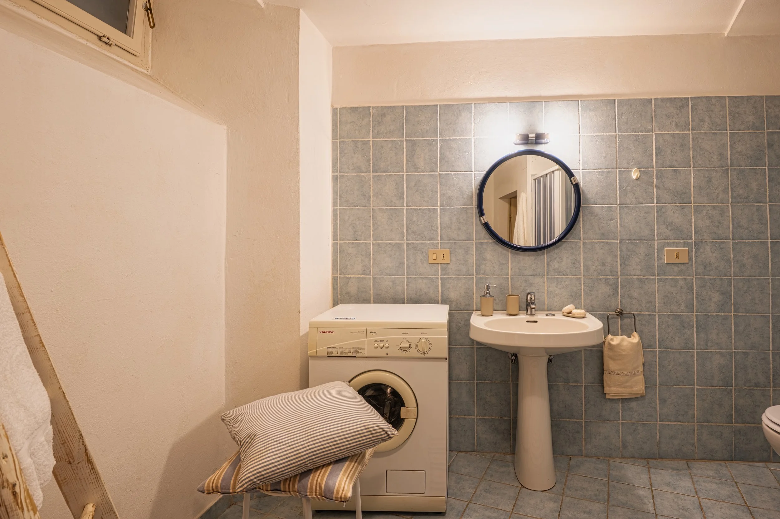 Bagno con lavatrice, lavabo con specchio rotondo e asciugamano su supporto. Pareti e pavimento rivestiti di piastrelle azzurre.