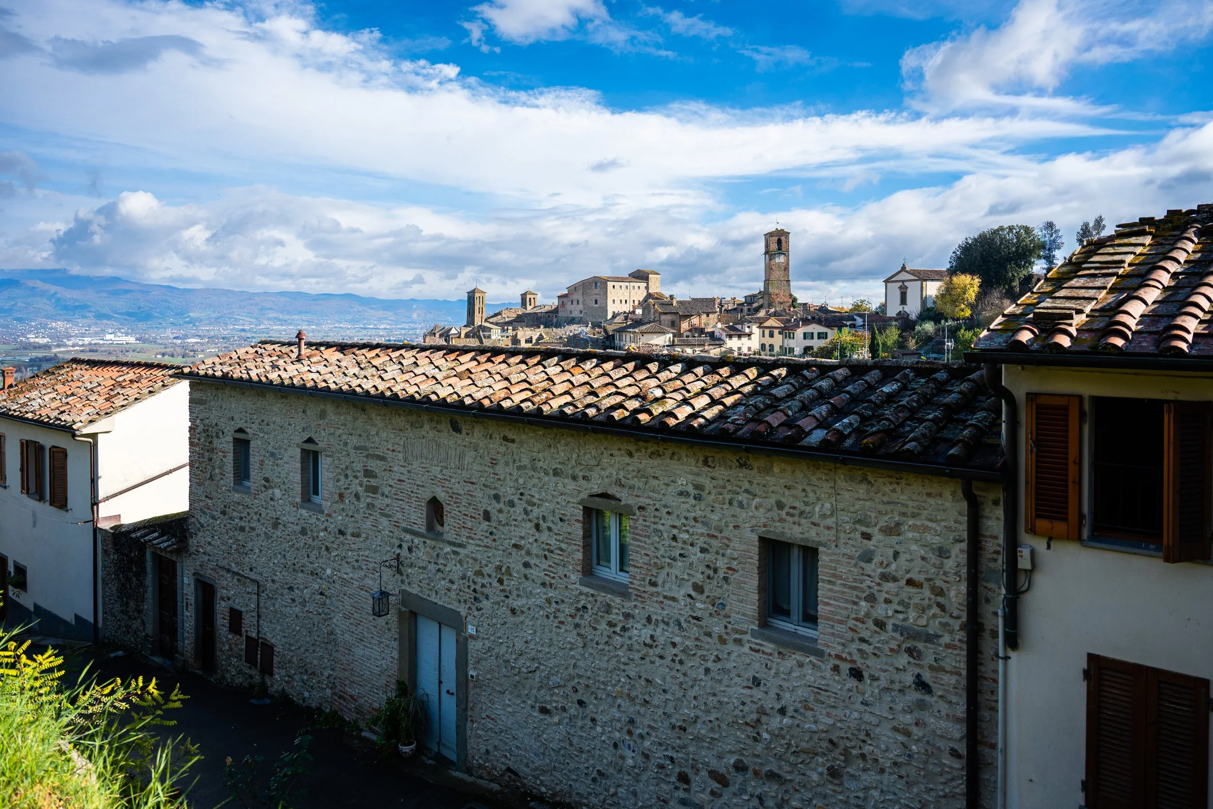 Vista di un paesino con case in pietra e tetti di terracotta, con un borgo medievale sopra, su una collina, sotto un cielo nuvoloso con aree di sole.