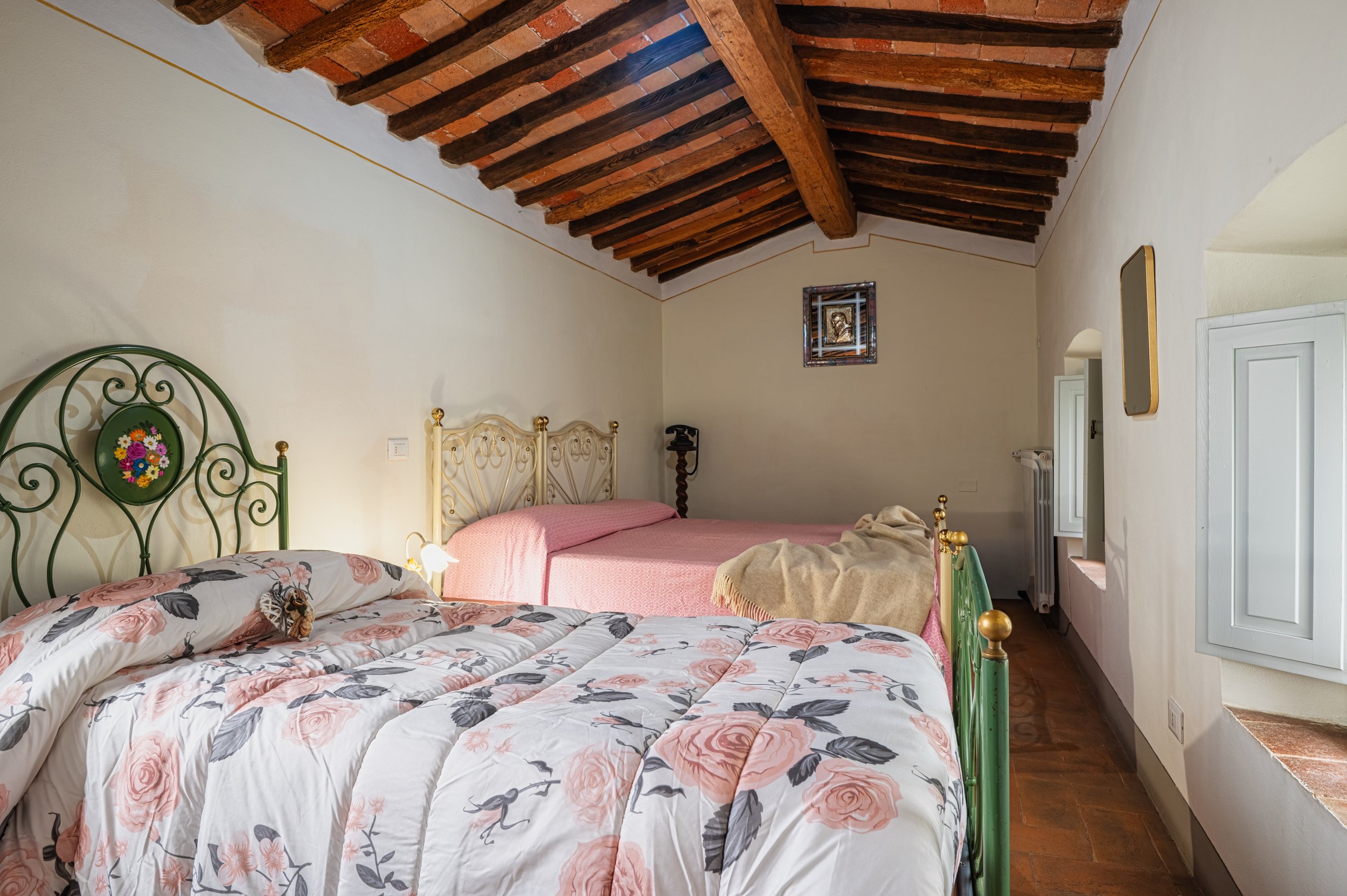 Camera da letto con soffitto in legno, due letti, uno con testa di ferro verde con decorazioni floreali e l'altro con testata in ferro bianco, coperte rosa e tessuto beige. Finestra con persiana bianca, pareti bianche e pavimento di piastrelle in ter