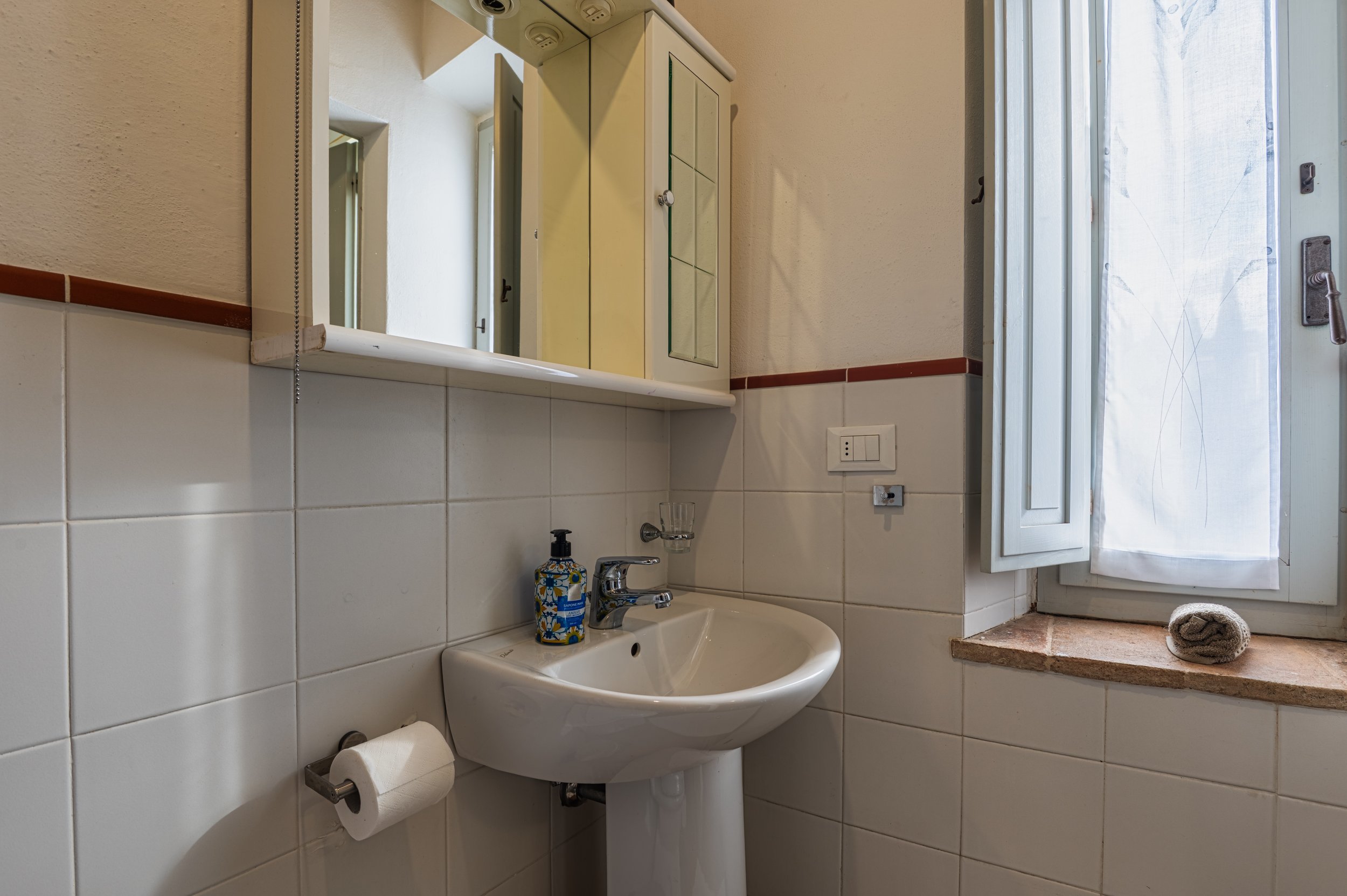 Bagno con lavabo, specchio, armadietto, finestra con tende e rotolo di carta igienica.