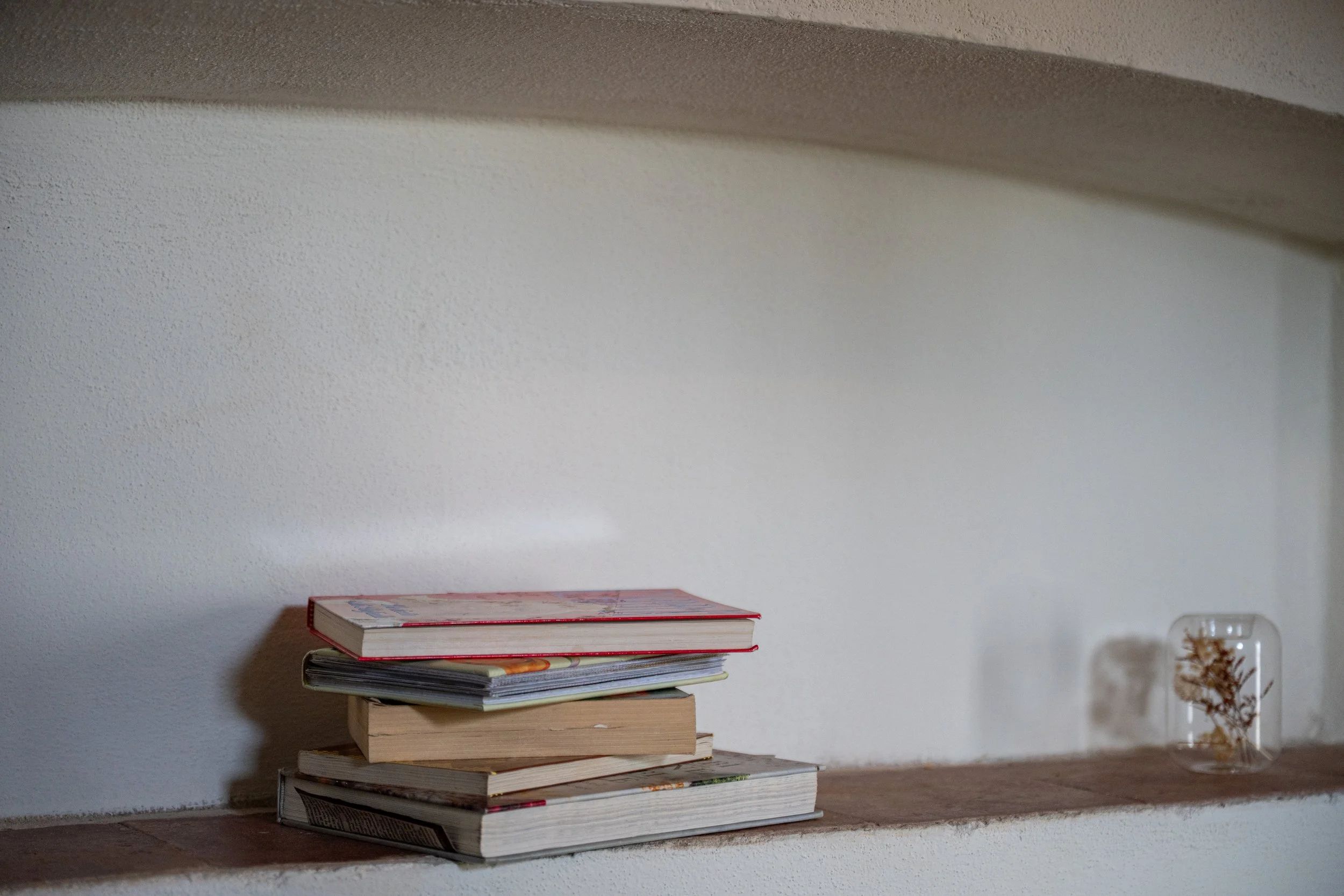 Pila di libri e riviste su una mensola di pietra accanto a un vaso con un fiore secco, su una parete bianca.
