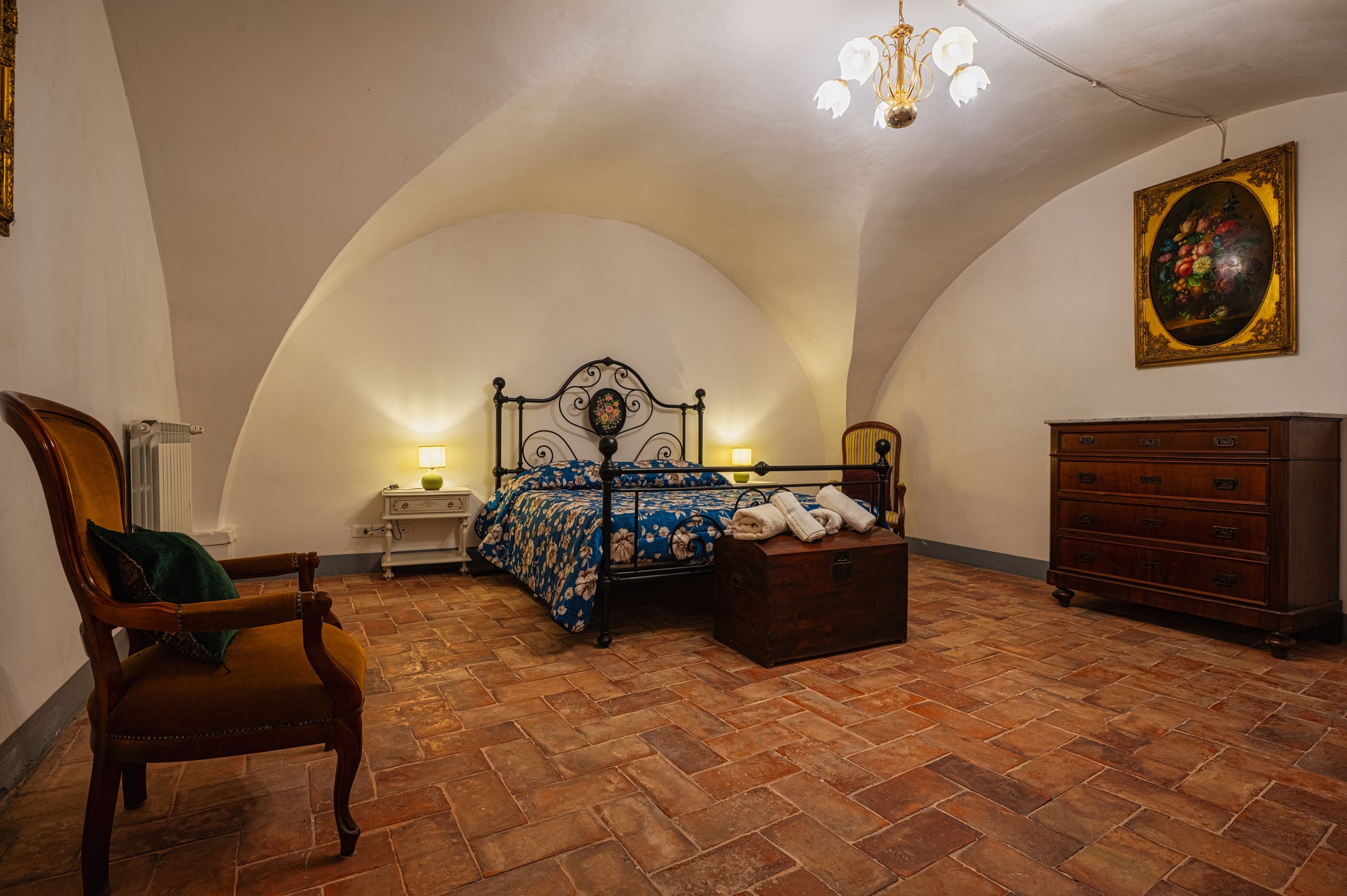 Una stanza da letto con pareti in pietra, letto in ferro battuto con coperta blu decorata con fiori, due lampade sui comodini, una sedia antica in legno, un taccuino con asciugamani e un grande mobile in legno. Sull'angolo destro, un quadro con una c