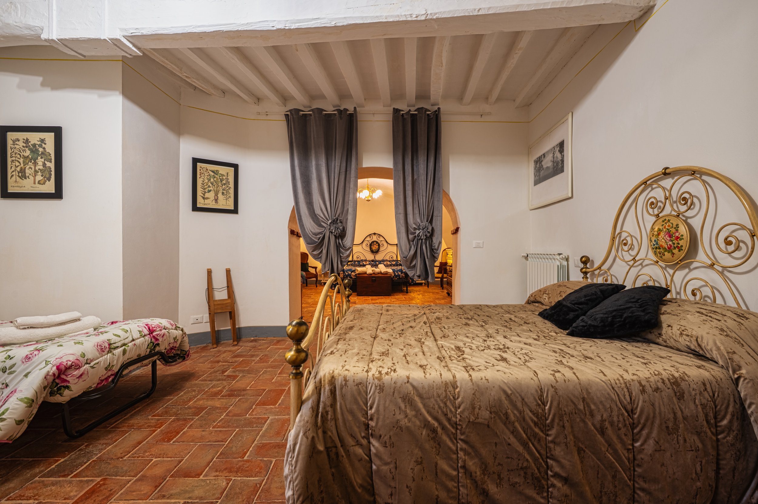 Camera da letto arredata con letto a baldacchino, cuscini neri, e decorazioni in stile vintage, con pareti bianche e pavimento in piastrelle rosse, e una porta aperta che mostra un'altra stanza con mobili d'epoca.