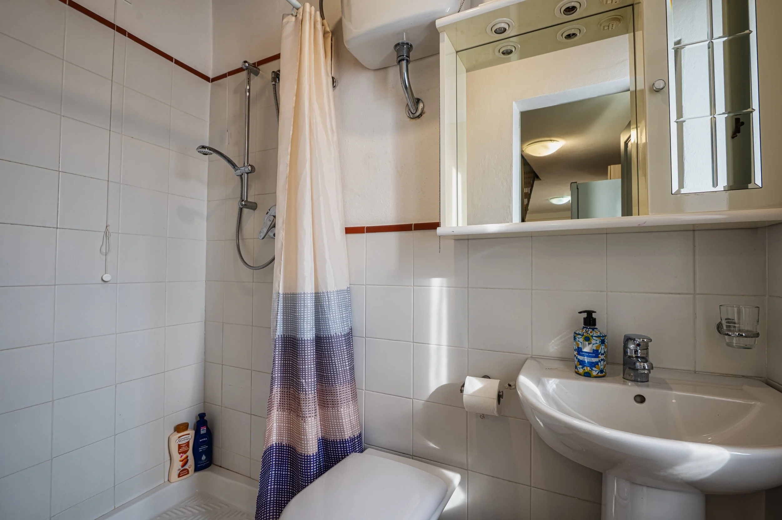 Bagno con doccia, tenda a righe blu, bianca e grigia, lavabo con dispenser di sapone, specchio e porta rotoli di carta igienica, rivestimenti a piastrelle bianche con fascia di piastrelle marroni.