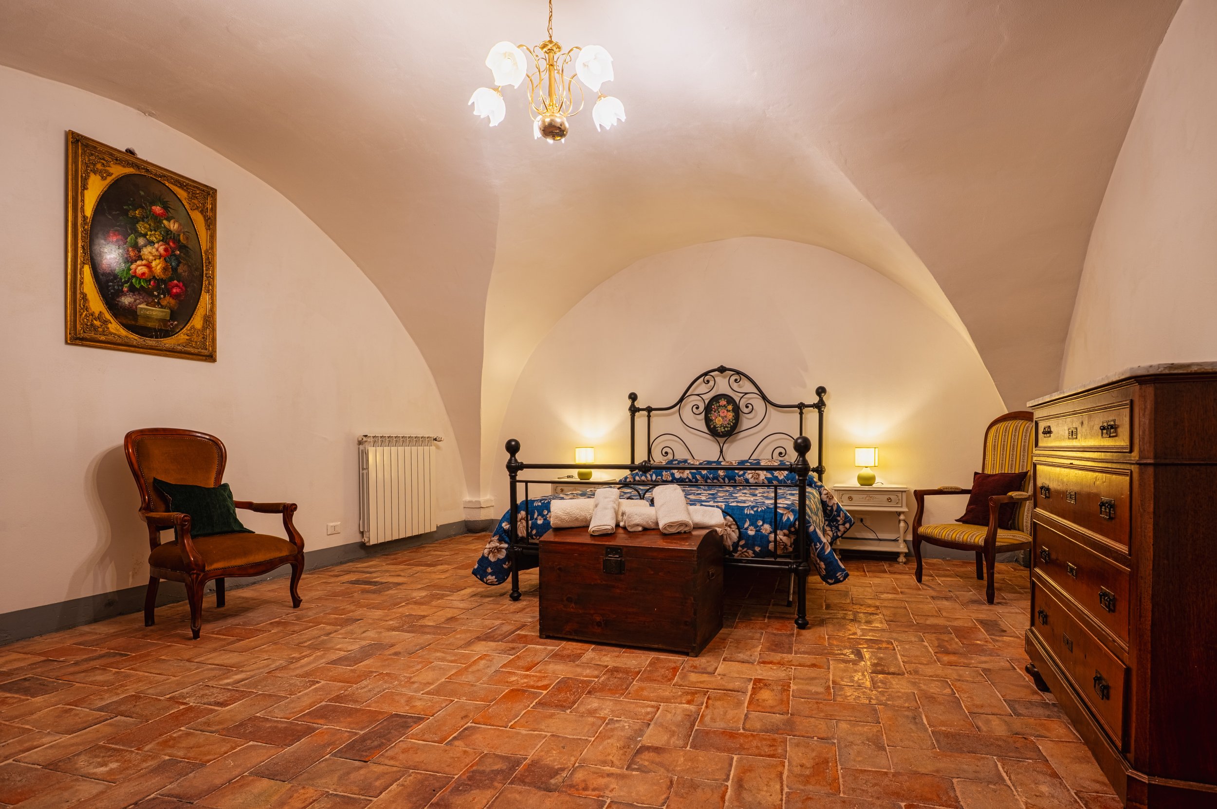 Camera da letto con pareti curve, pavimento in piastrelle di terracotta, letto con testiera in ferro battuto con copricostiera blu con fiori, quattro asciugamani bianchi sul letto, due lampade da comodino accese, armadio in legno scuro, sedie in legn