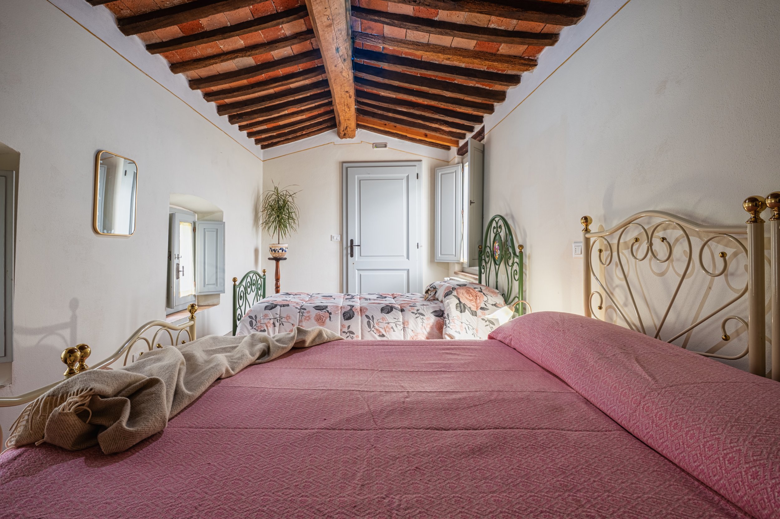 Camera con due letti, uno con copriletto rosa e l'altro con copriletto floreale, pareti bianche, finestre con persiane azzurre, soffitto con travi in legno a vista e piante.