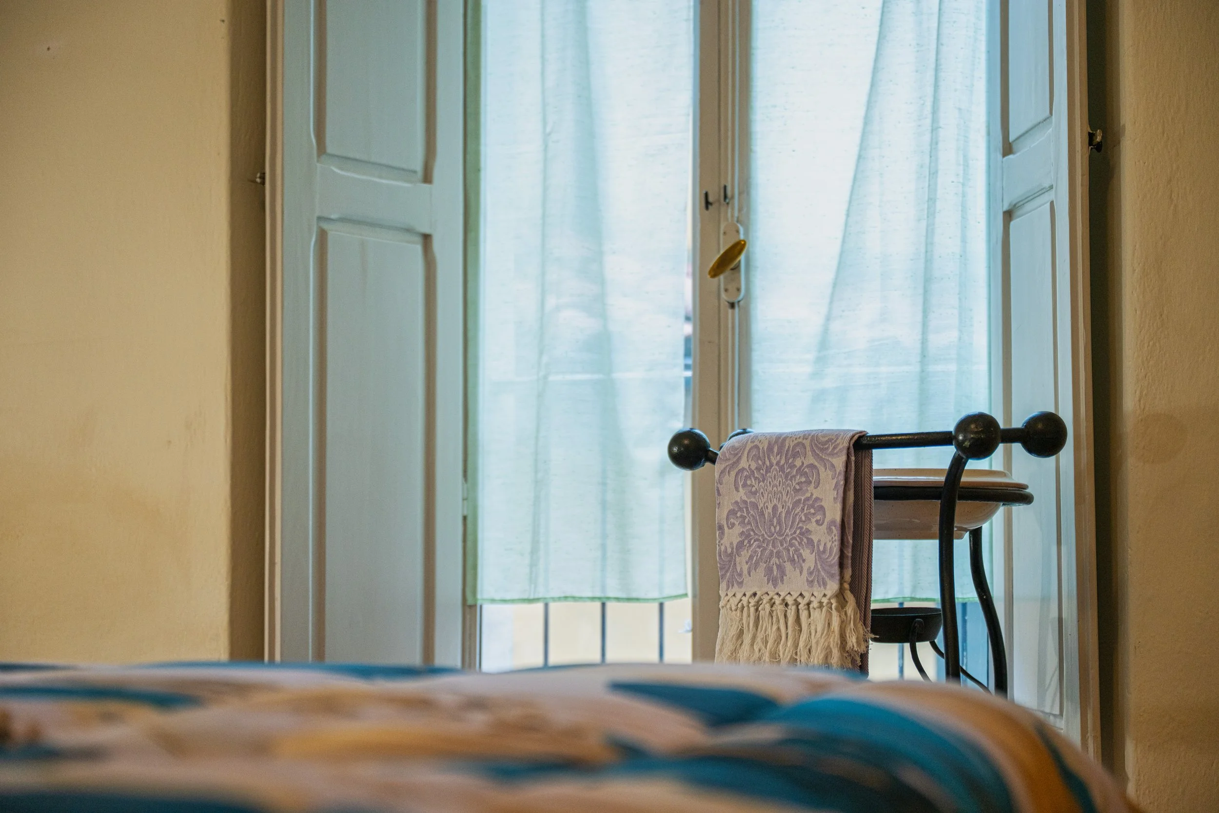 Vista di una stanza con una finestra aperta con tende azzurre, una sedia con un asciugamano appoggiato e di fronte una parete gialla.