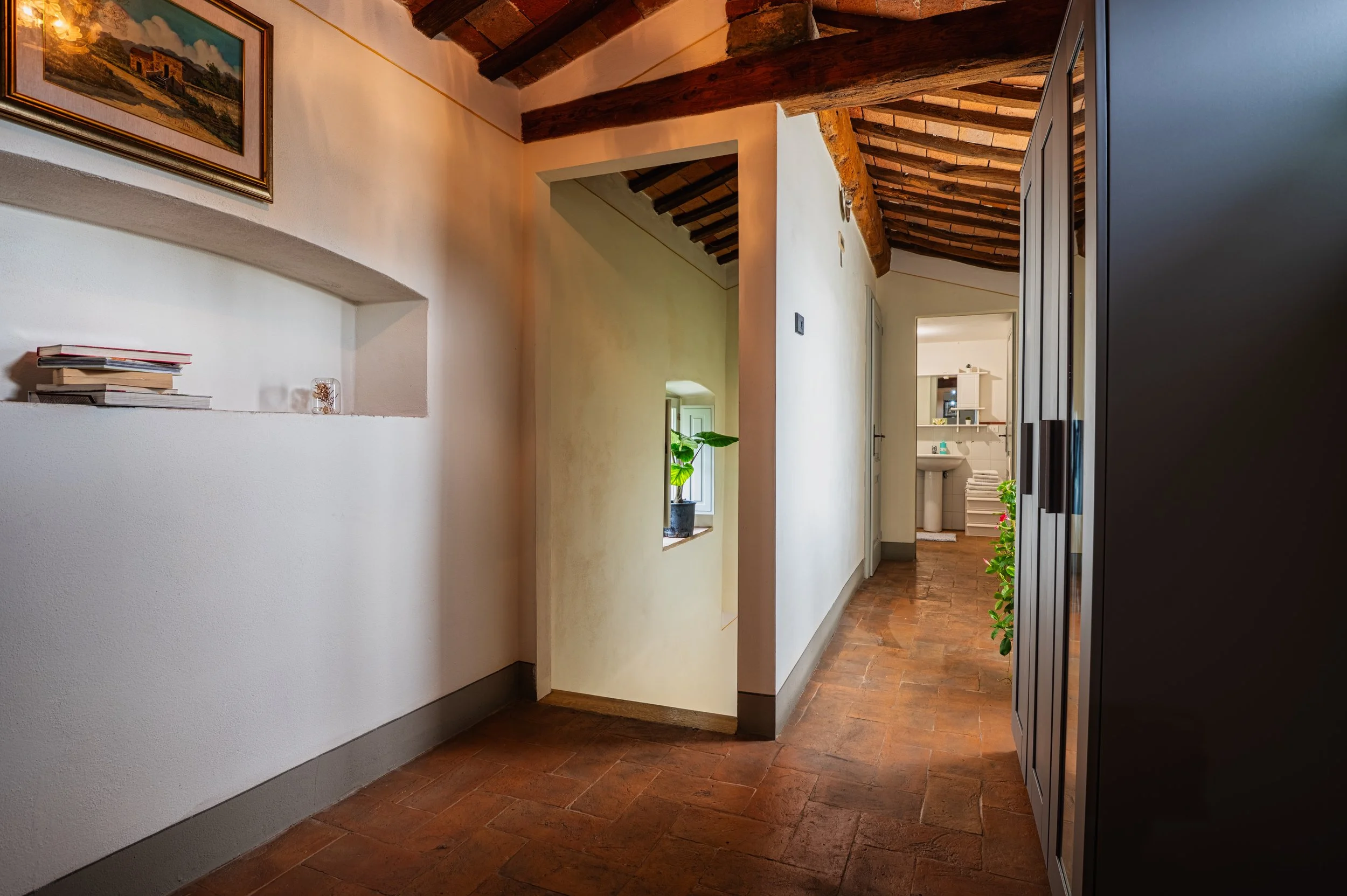 Interno di una casa con pavimento in terracotta, pareti bianche, soffitto con travi in legno a vista, porta aperta che rivela un bagno con lavabo, monstersca e scaffali.