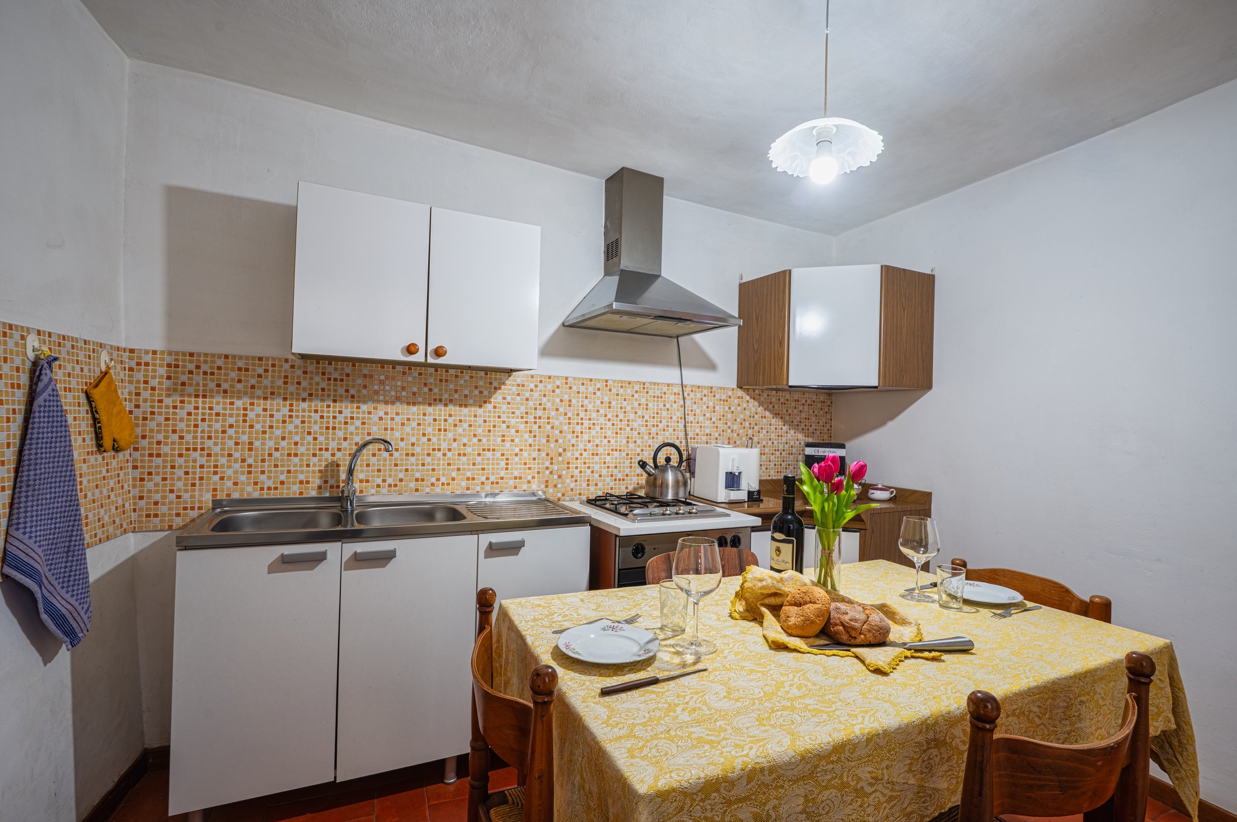 Cucina con tavolo apparecchiato con pane, vino, bicchieri e fiori, e cucina con armadietti bianchi e forno a gas.