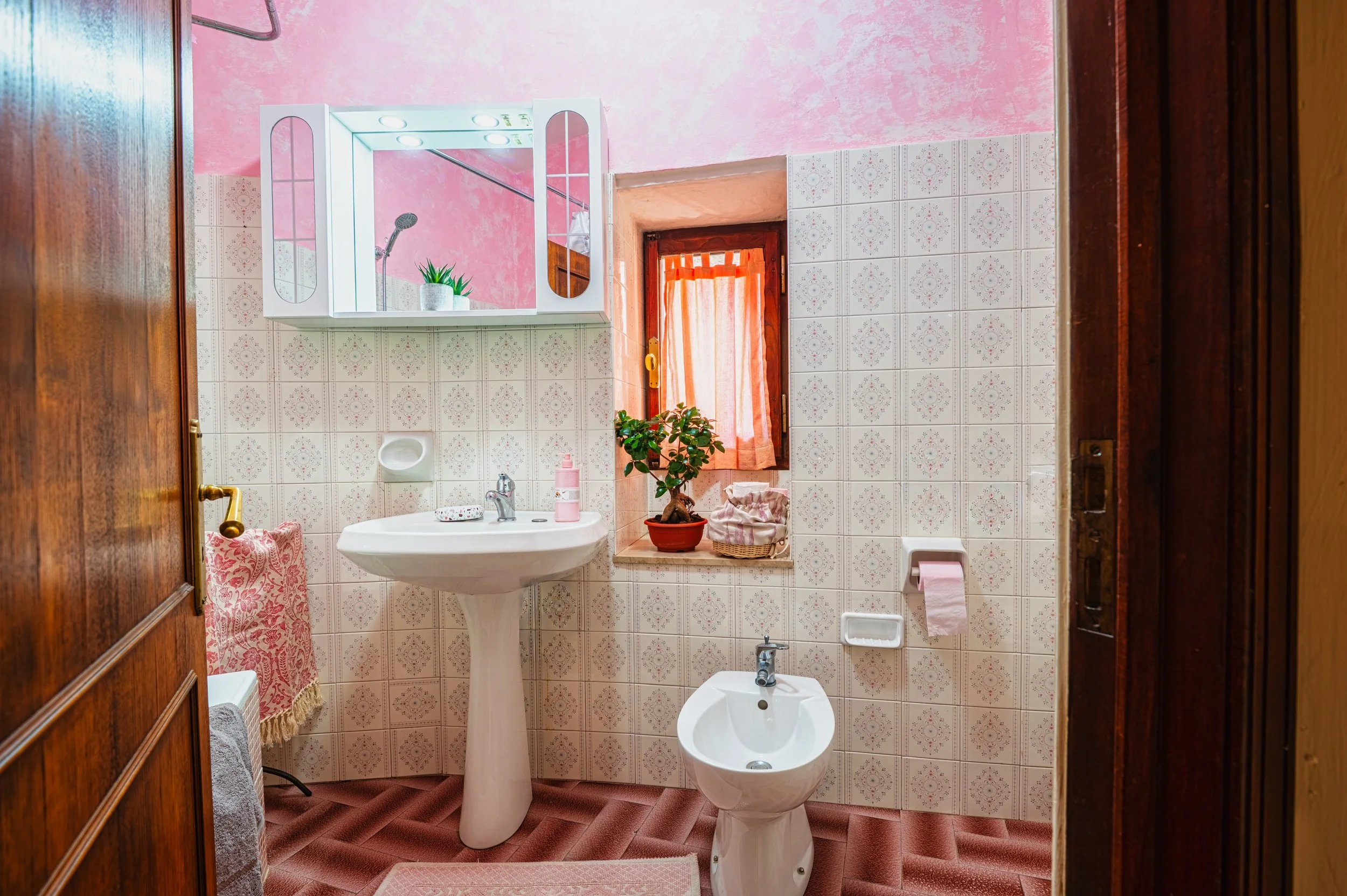 Bagno con pareti rivestite di piastrelle beige con motivi rosa, lavabo con specchio e luci sopra, bidet, finestra con tende rosa, piante e accessori bagno rosa, pavimento in piastrelle rosse.