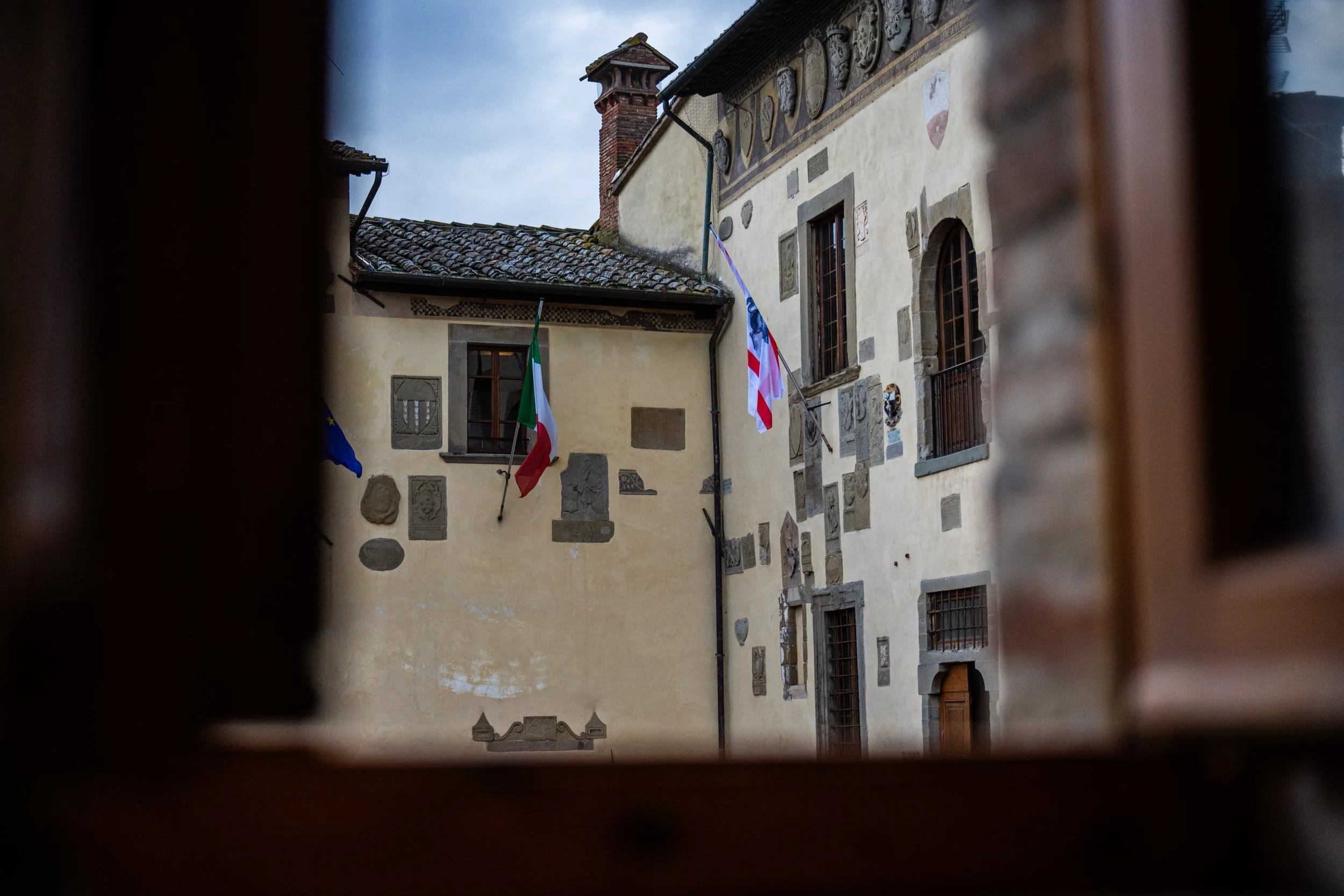 Vista di un cortile con pareti di un edificio in pietra e affreschi, con tre bandiere appese: quella dell'Unione Europea, quella italiana e quella degli Stati Uniti.
