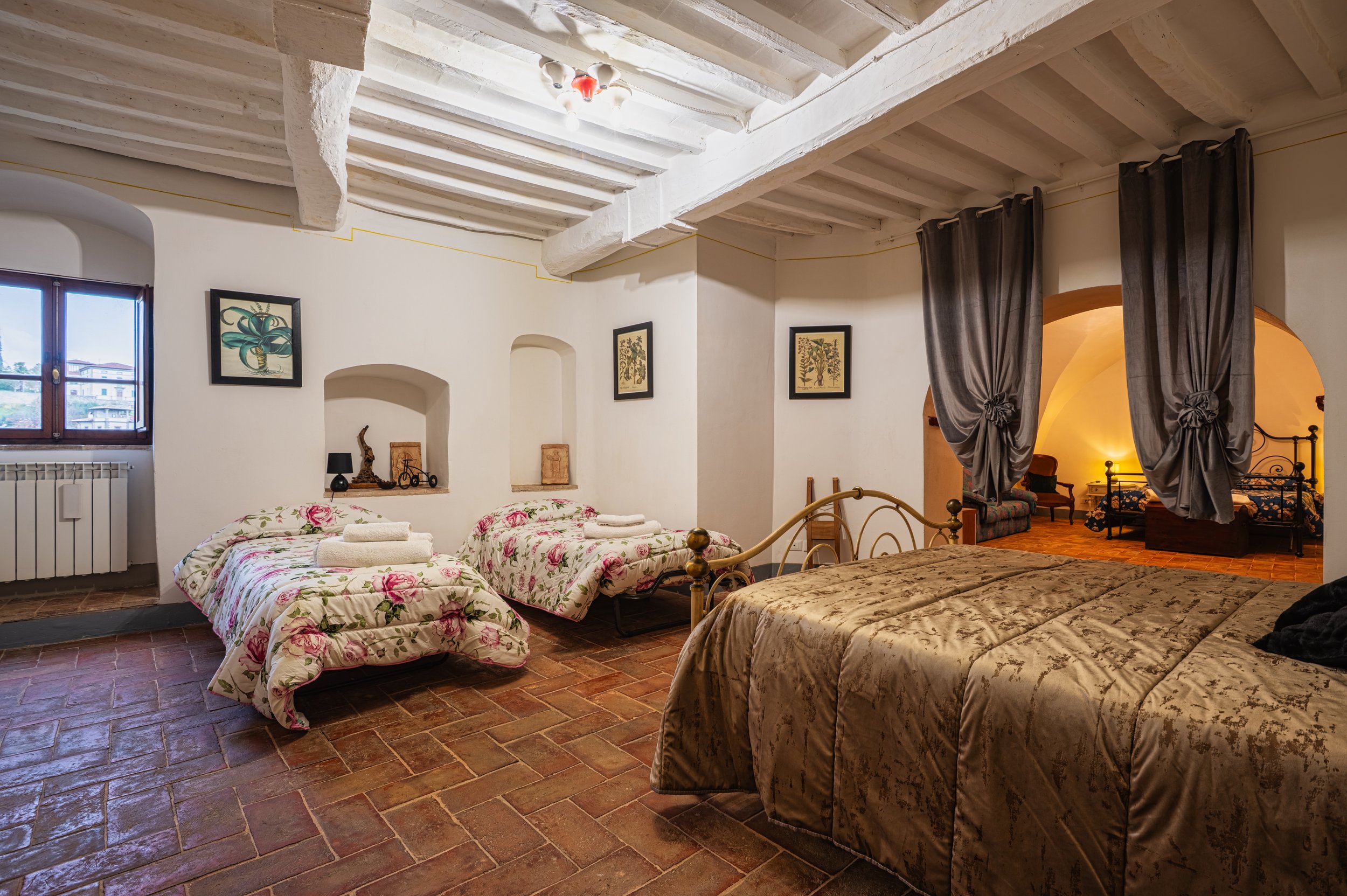 Camera con due letti singoli con copripiumini floreali e un grande letto matrimoniale con copriletto marrone. Pareti bigrate con nicchie e quadri. Finestra con vista su edifici. Tappeto di mattoni e soffitto con travi di legno bianco.