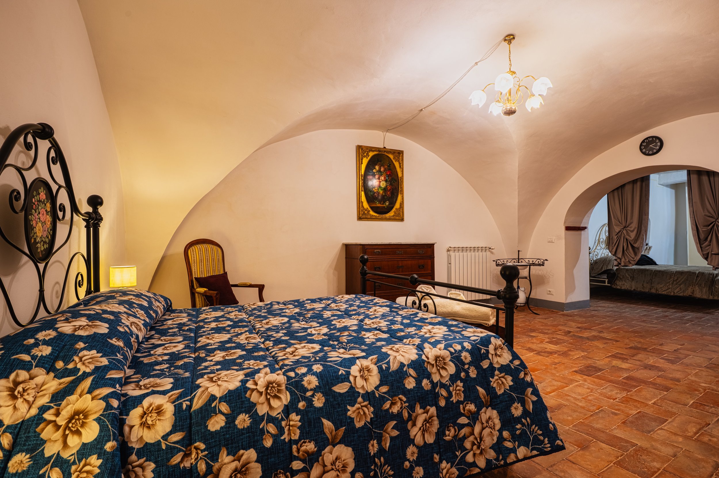 Camera da letto arredata in stile classico con soffitto con arcate, grandi finestre con tende di velluto, pavimento in piastrelle di terracotta, letto con copriletto a fiori, dipinto floreale, lampada accesa, mobili in legno e sedie vintage.