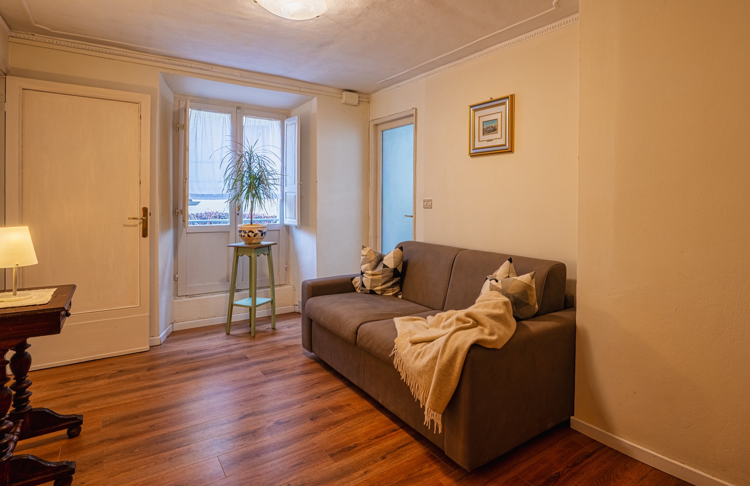 Soggiorno con divano marrone, cuscini decorativi, quadro alle pareti, finestra con tende neutre, lampada e tavolo in legno.