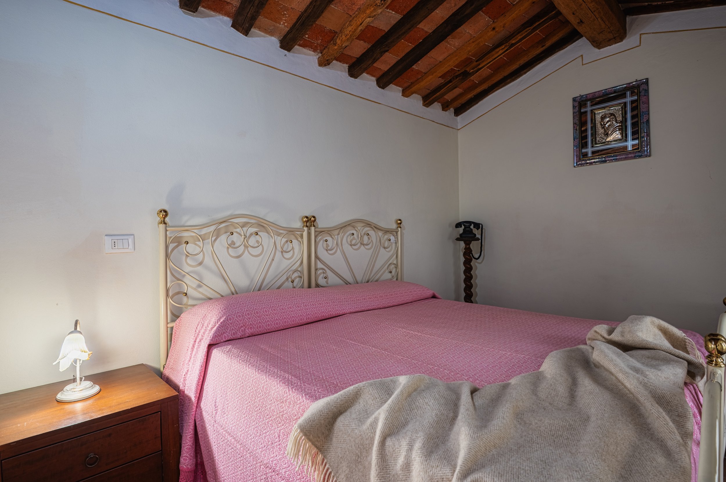 Camera da letto con letto matrimoniale con testiera in metallo decorato, coperta rosa, e coperta beige svolazzante. Accanto al letto, comodino in legno con piccola lampada, e sullo sfondo, pareti bianche, soffitto con travi in legno, e quadro con cor