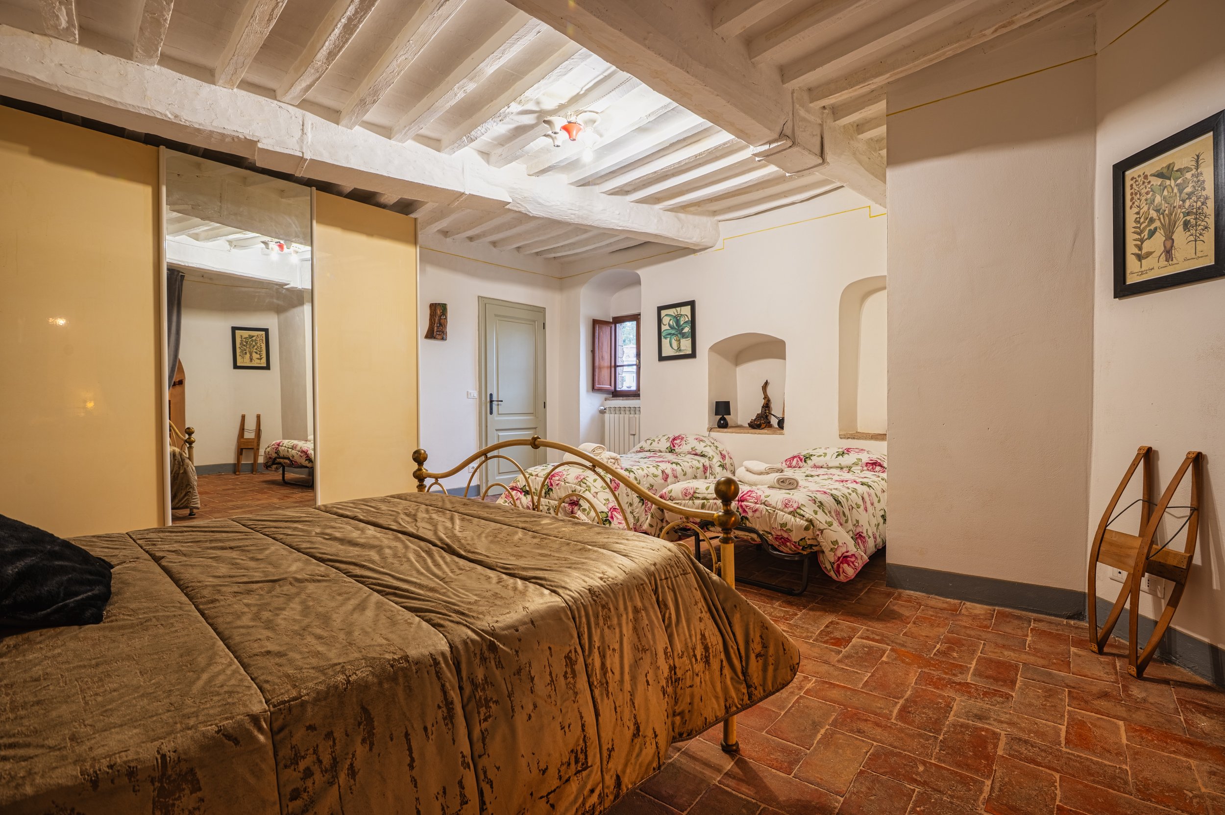 Camera con due letti singoli con copriletti floreali, letto matrimoniale con coperta marrone, specchio e pareti decorate con quadri, pavimento in piastrelle di terracotta, mobili in legno, finestra aperta con vista sulla strada