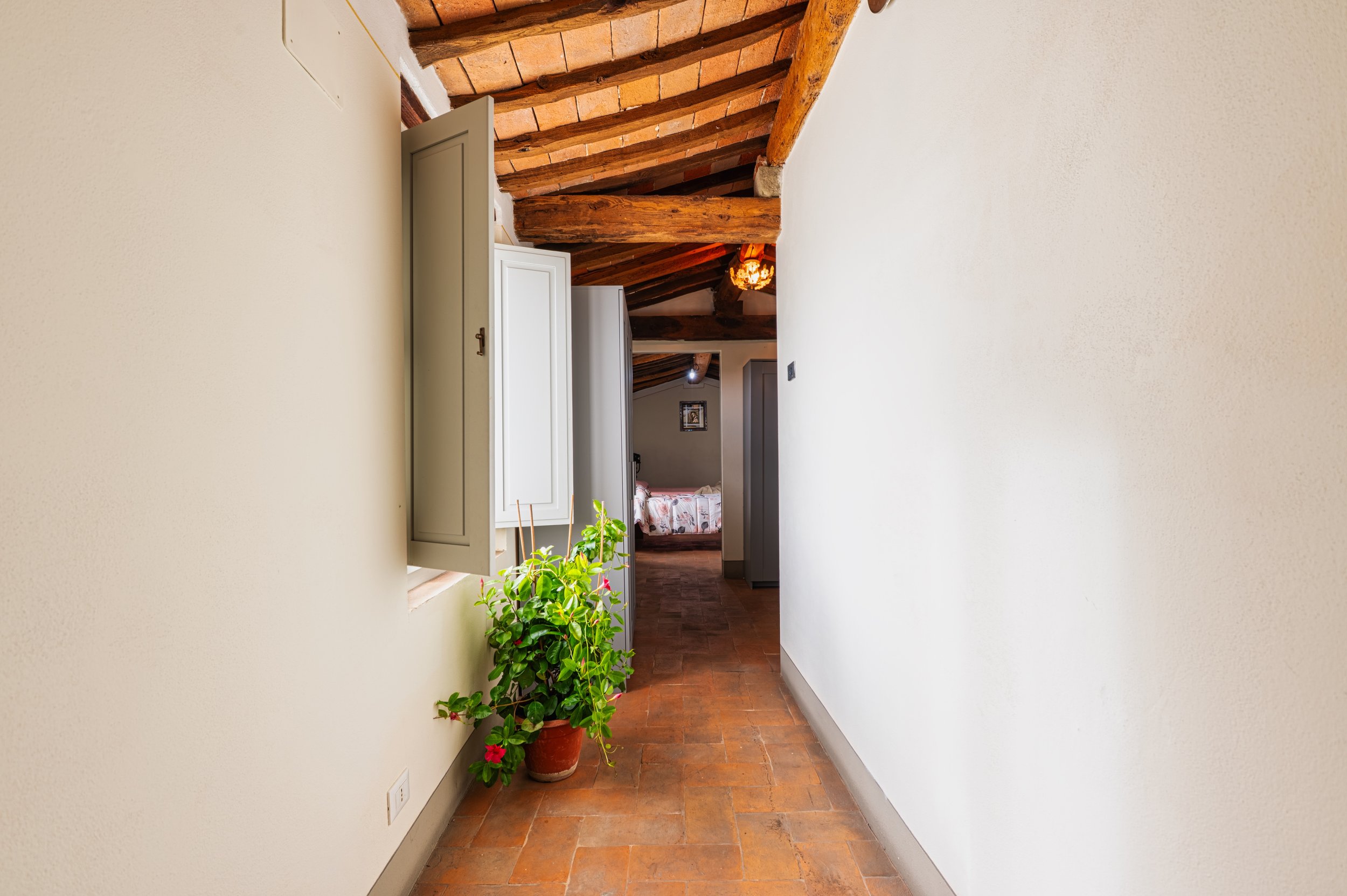 Corridoio in una casa rustica con soffitto di travi di legno e terracotta, finestra aperta con fiori in vaso, vista verso una stanza con letto.