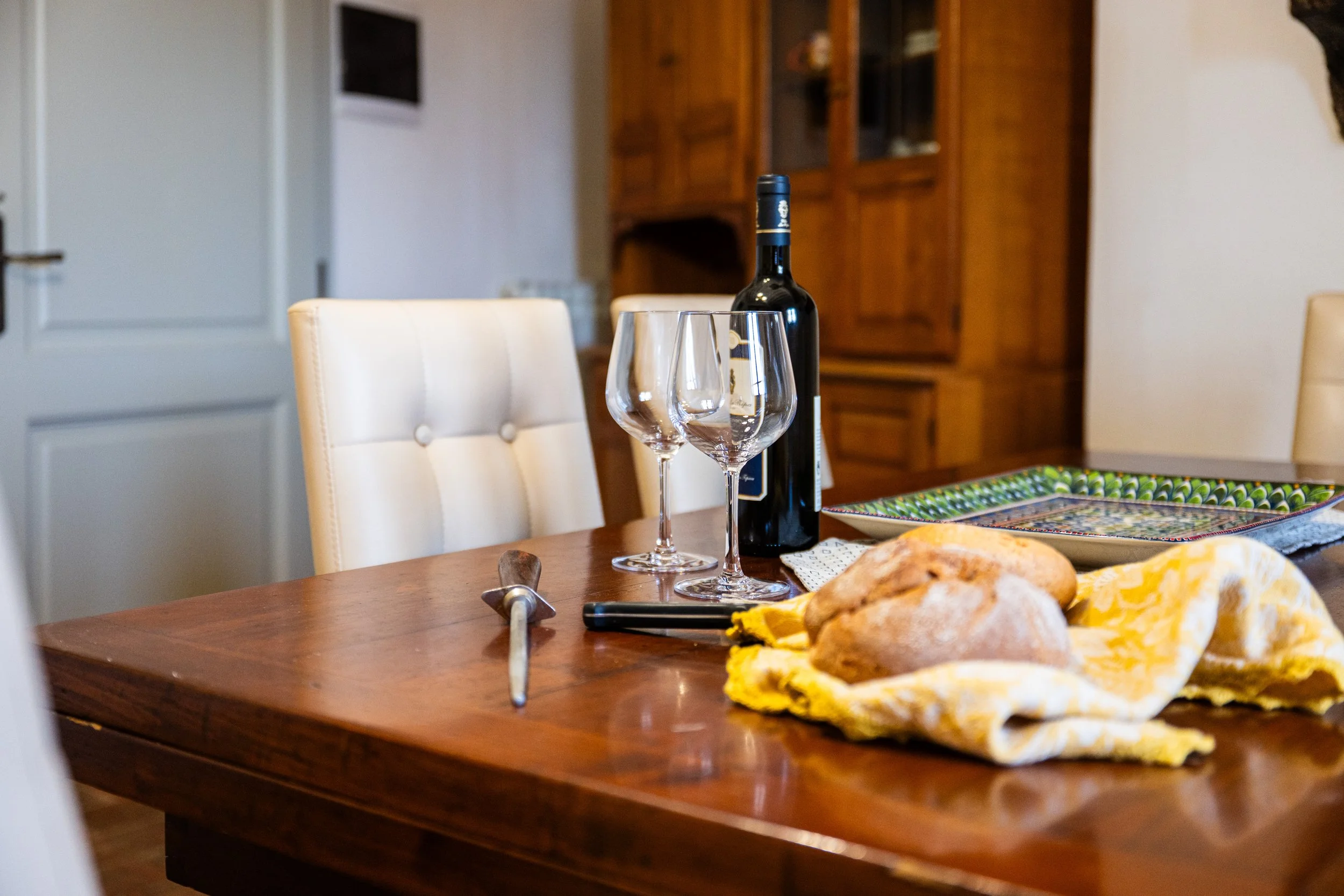 Tavolo apparecchiato con una bottiglia di vino, due calici vuoti, pane su un panno giallo, un coltello e una posata, in una stanza con sedie chiare e mobili in legno.