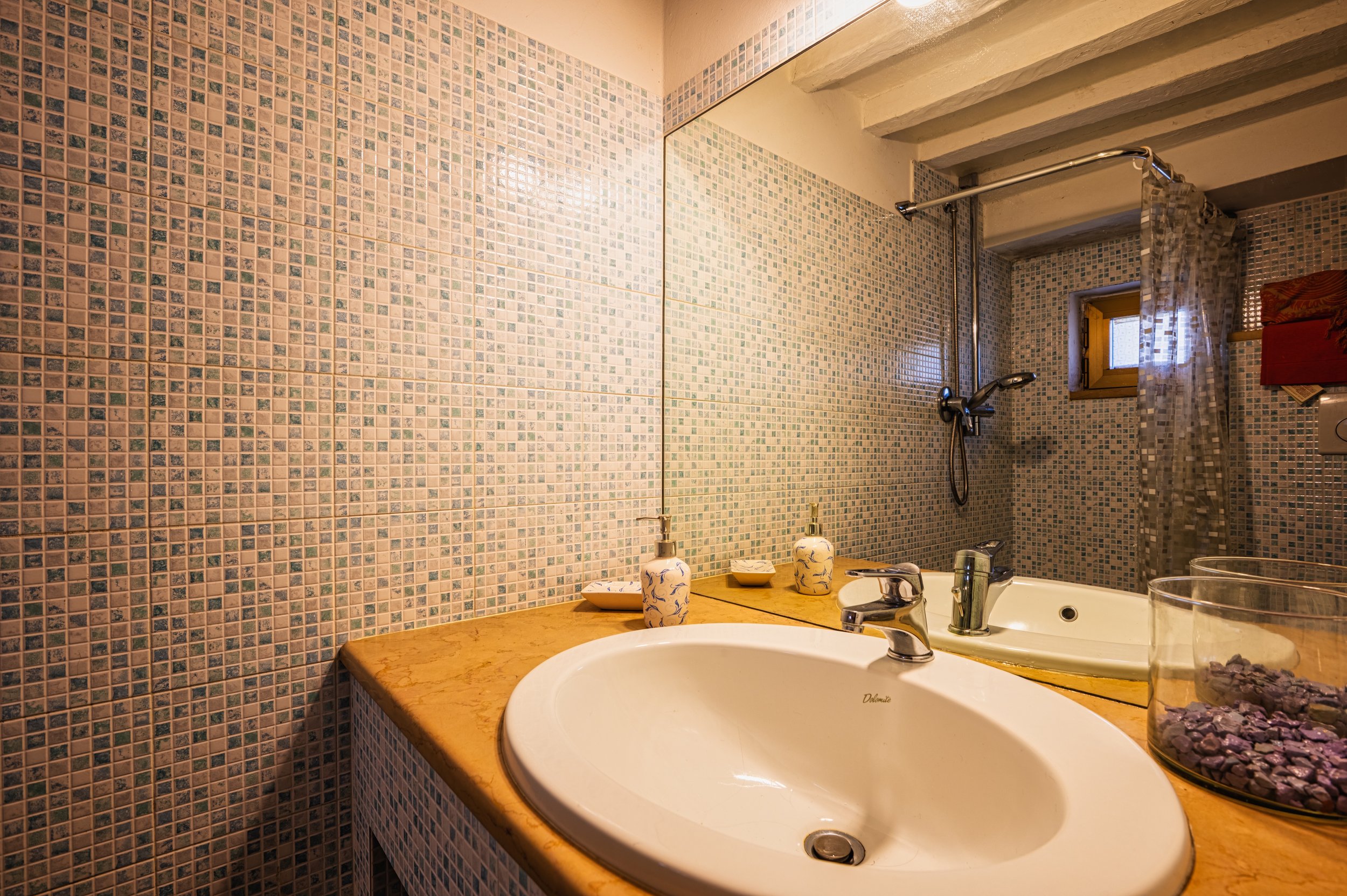 Bagno con lavabo in ceramica, specchio grande, muro rivestito con piastrelle color beige e azzurre, finestra con tenda trasparente, doccia con cortina, accessori per il bagno e ciotola con pietre decorative.