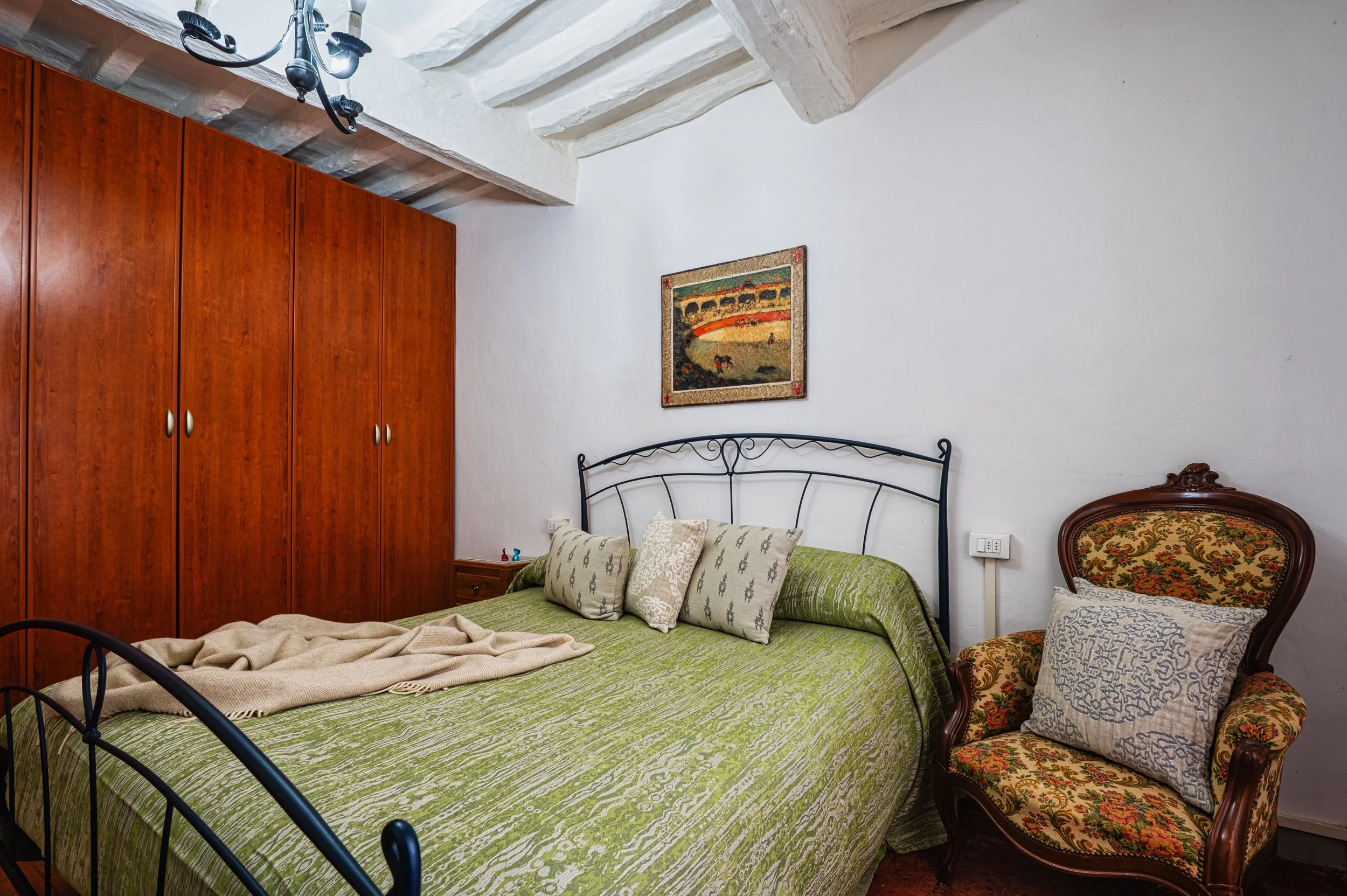 Camera da letto con letto a baldacchino con copriletto verde, alcuni cuscini, un armadio in legno scuro, una sedia antica con rivestimento damascato, pareti bianche e un quadro appeso alla parete.