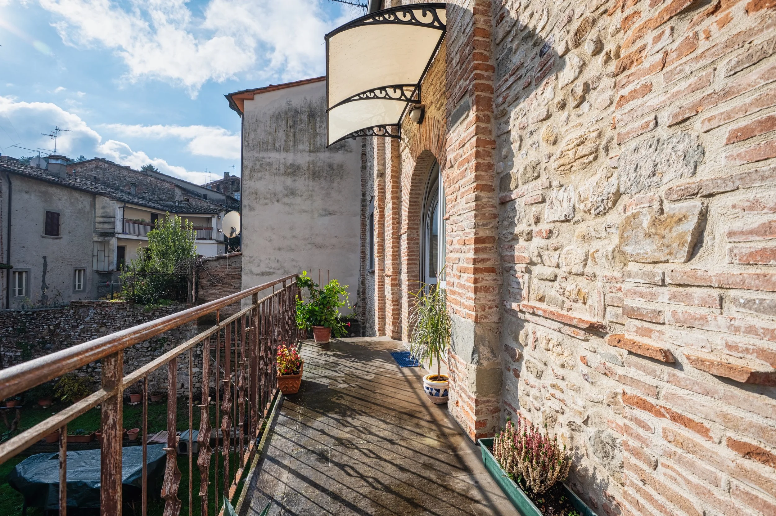 Balcone di una casa con piantine in vasi, pareti in mattoni e pietra, sotto un cielo con nuvole. 