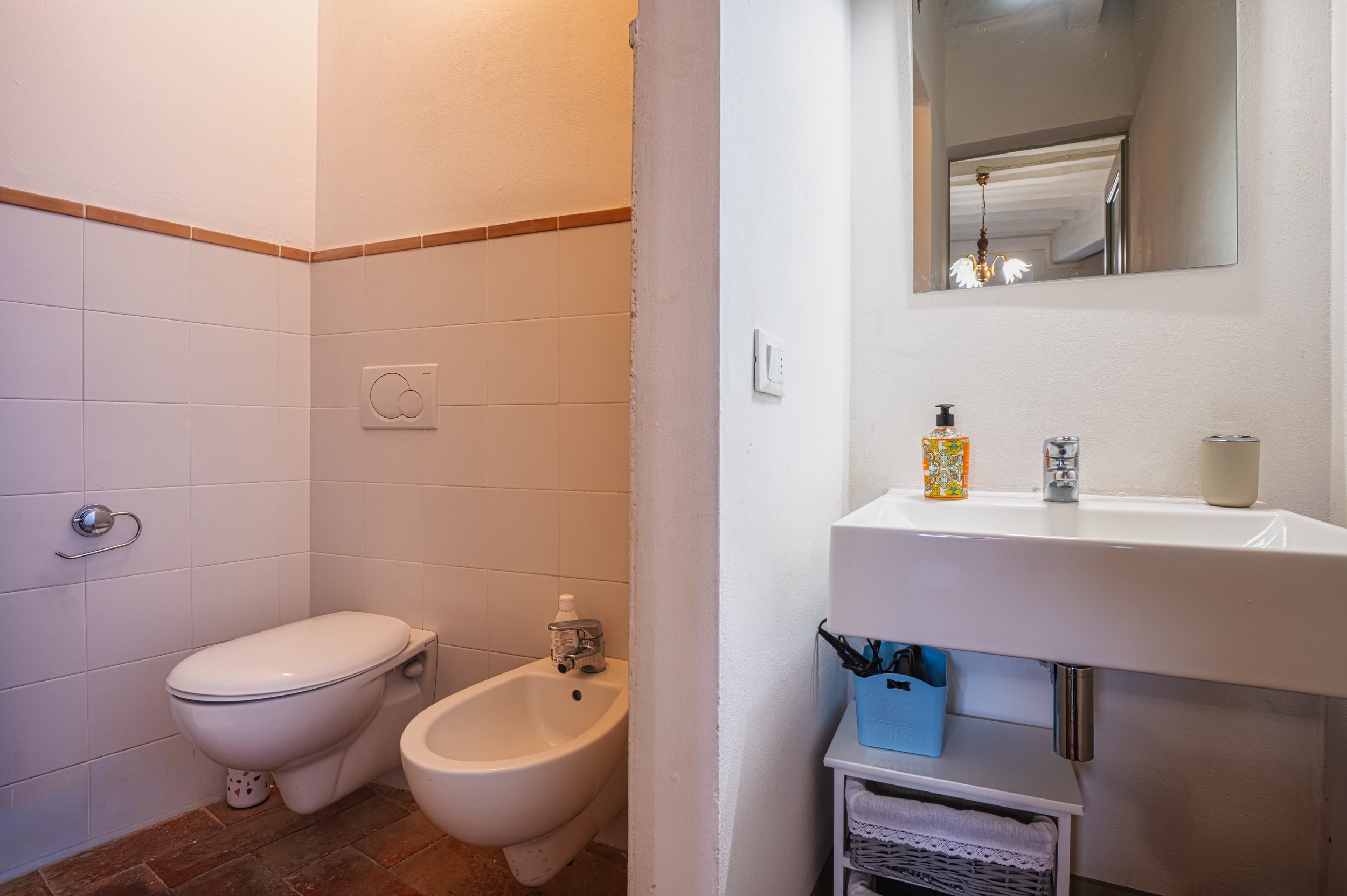 Bagno con wc e bidet vicino, lavandino con specchio sopra, sapone e portaspazzolino, sotto un mobile con scaffale, pareti bianche e piastrelle beige con lista marrone.