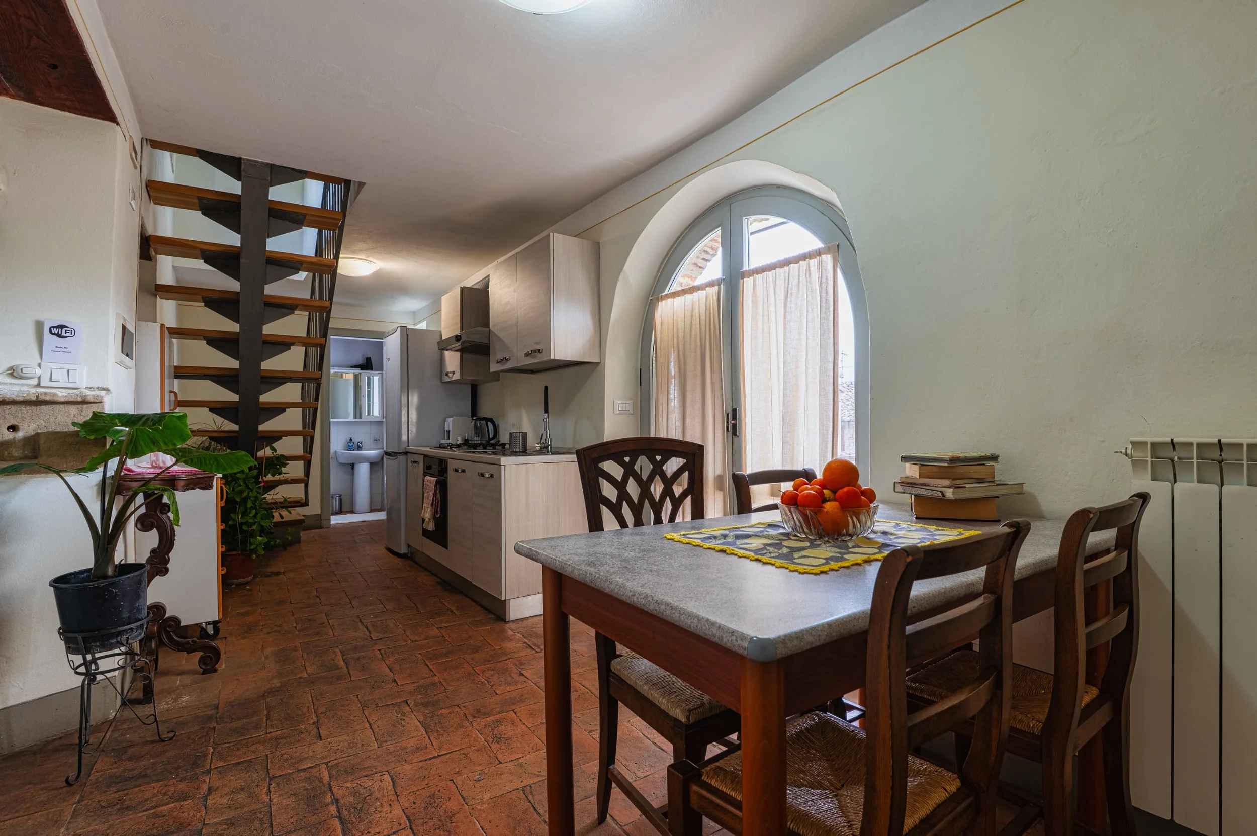 Angolo cucina e pranzo di una casa con tavolo, sedie, cucina compatta, scale a chiocciola, piante verdi, finestra con tende beige, e pavimento in piastrelle rosse.