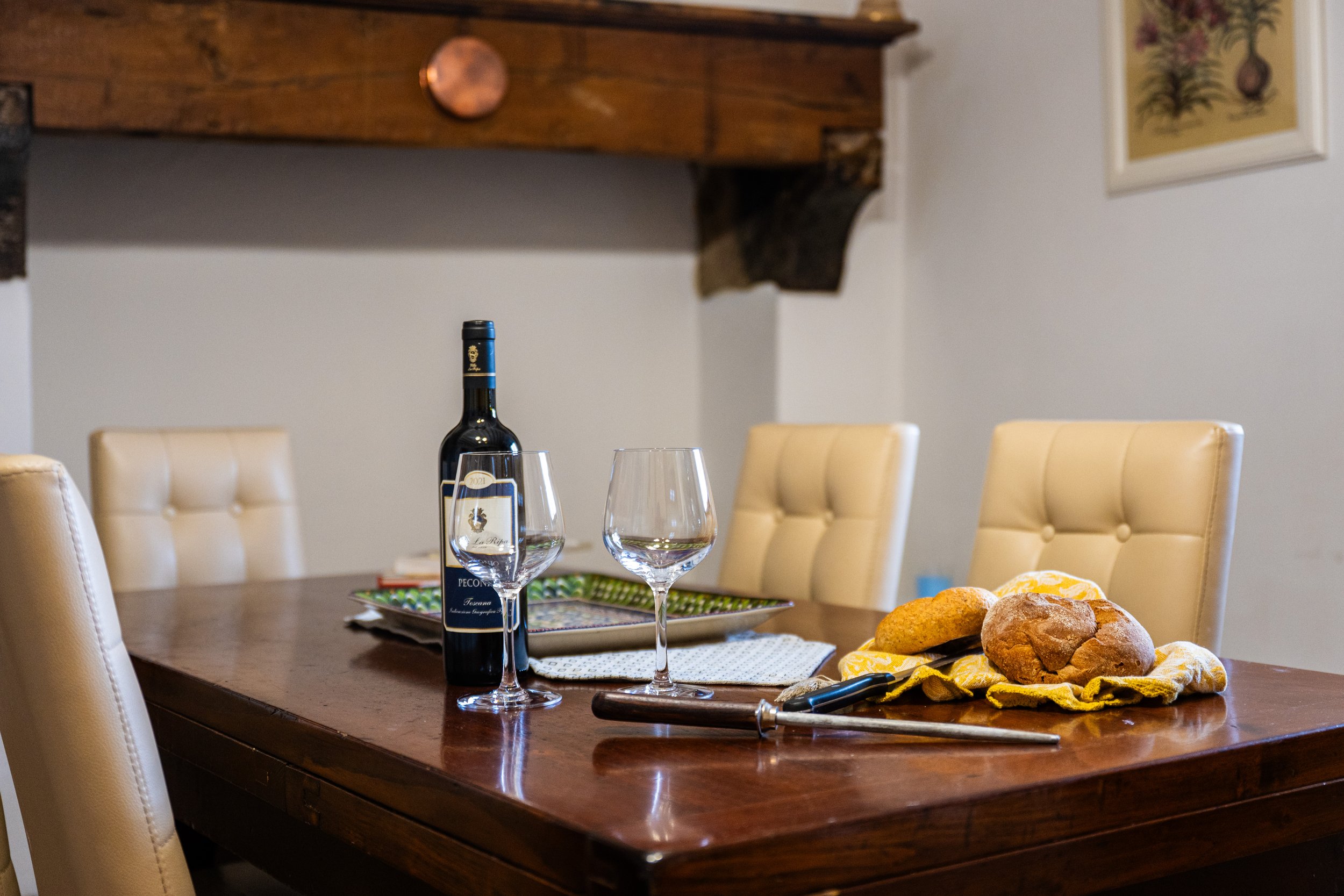 Tavola di legno con una bottiglia di vino, due bicchieri vuoti, pane e focaccia su un tovagliolo giallo e utensili da cucina, in una stanza con sedie imbottite beige e pareti decorate.