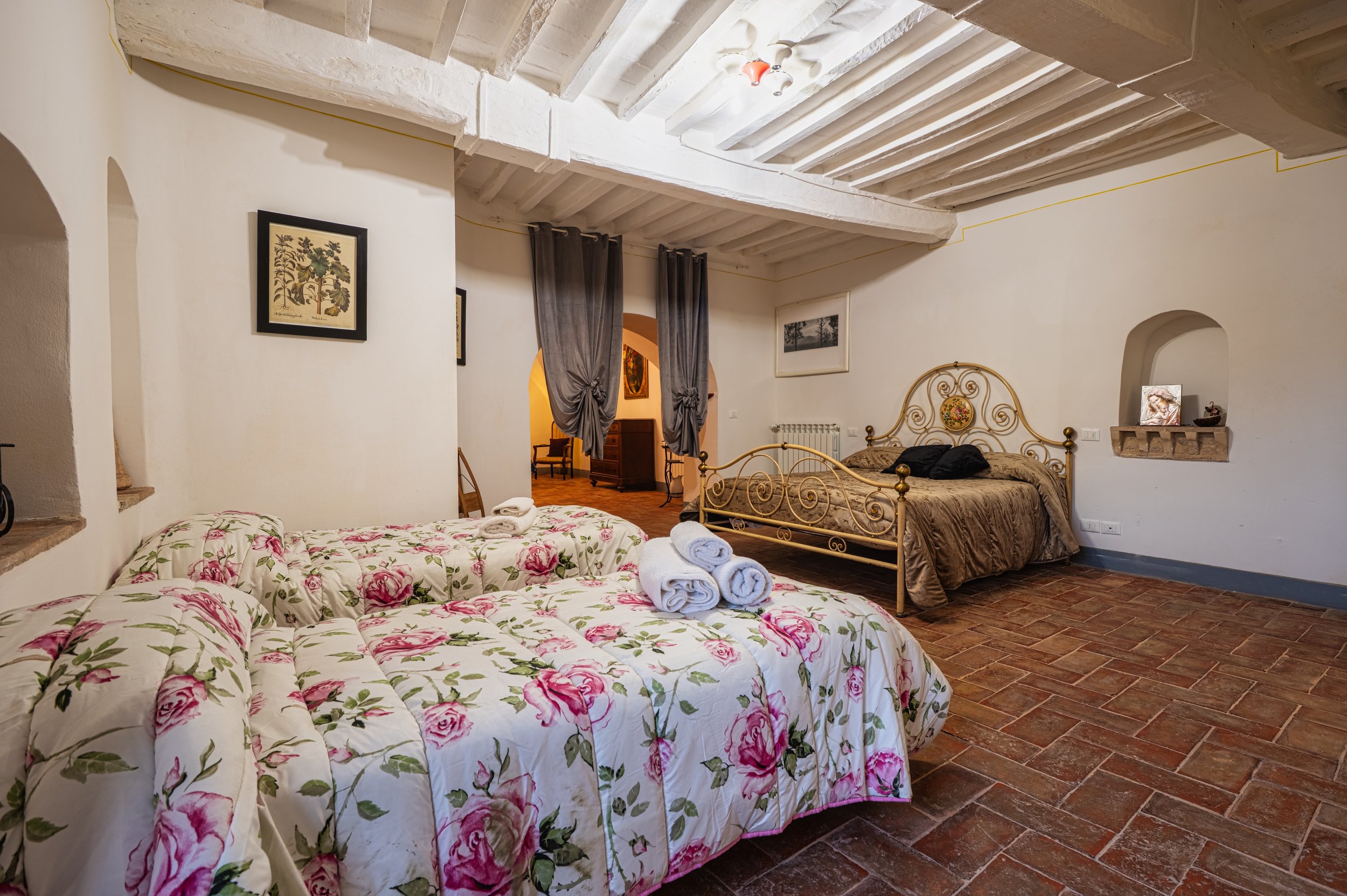 Camera da letto con due letti, uno con copriletto floreale e l'altro con copriletto marrone, decorata in stile rustico, con soffitto a travi bianche e pareti bianche. Ci sono guiding di tende grigie e quadri alle pareti.