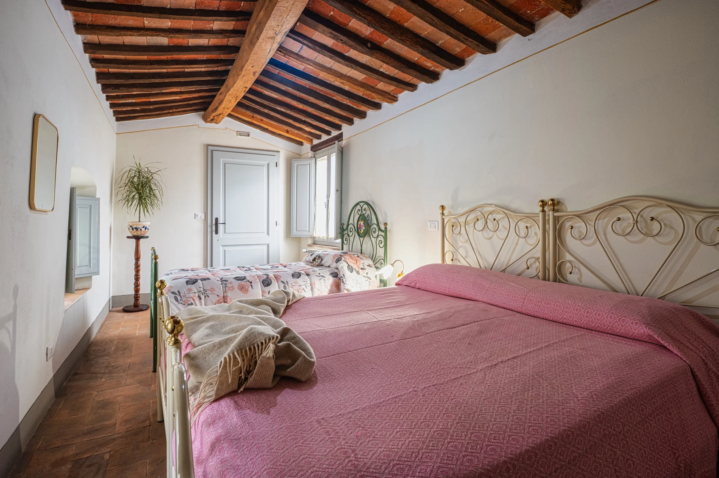 Camera da letto con due letti, uno con copertura rosa e l'altro con copertura floreale, soffitta con travi in legno a vista, finestra aperta, pareti bianche e pavimento di piastrelle in terracotta.
