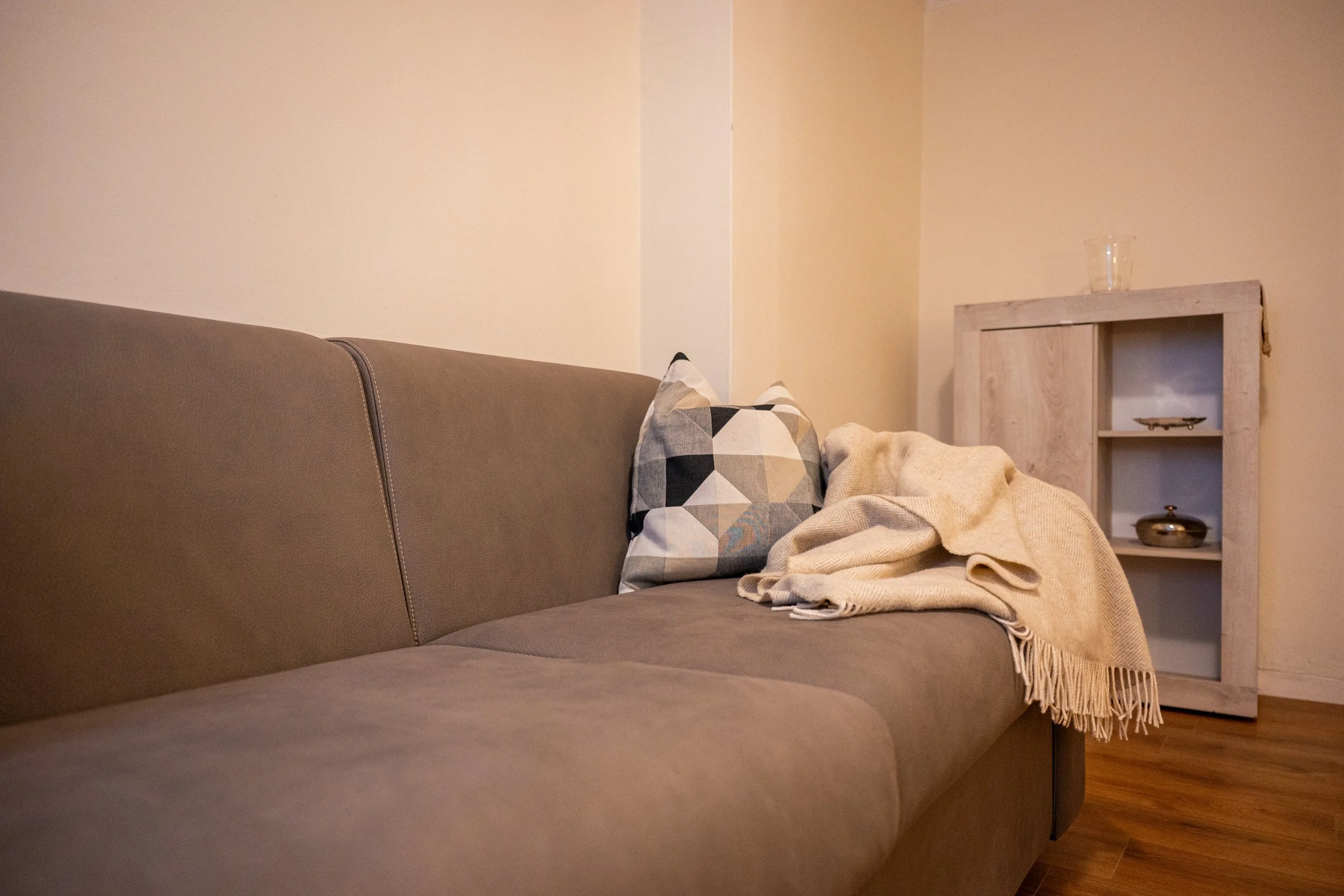 Soggiorno con divano marrone, cuscino con motivi geometrici, coperta beige, mobile in legno con oggetti decorativi, pareti beige, pavimento in legno.