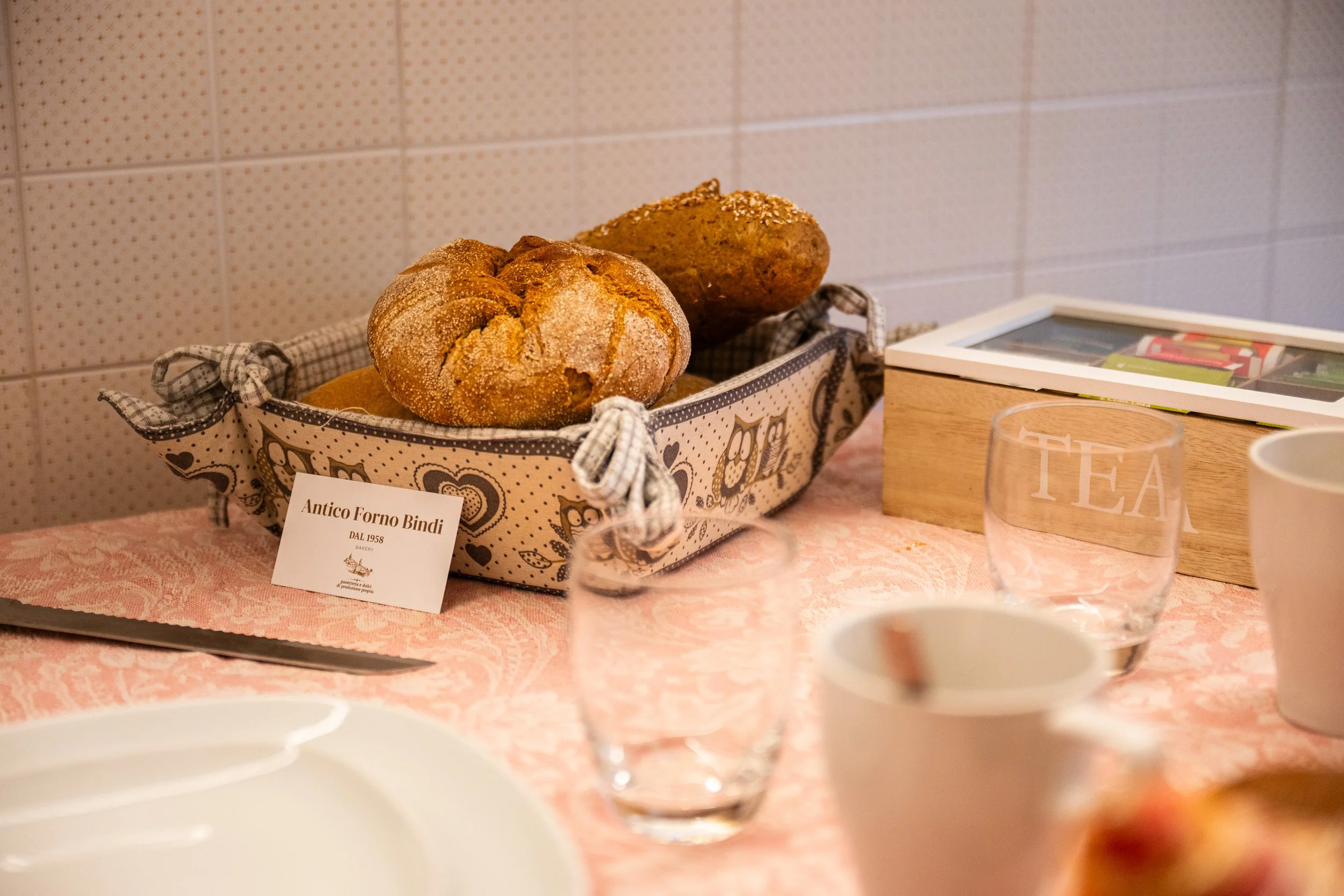 Pane e una torta in una cesta decorata, sul tavolo appare anche una tazza e due bicchieri con scritto 'TEA'.
