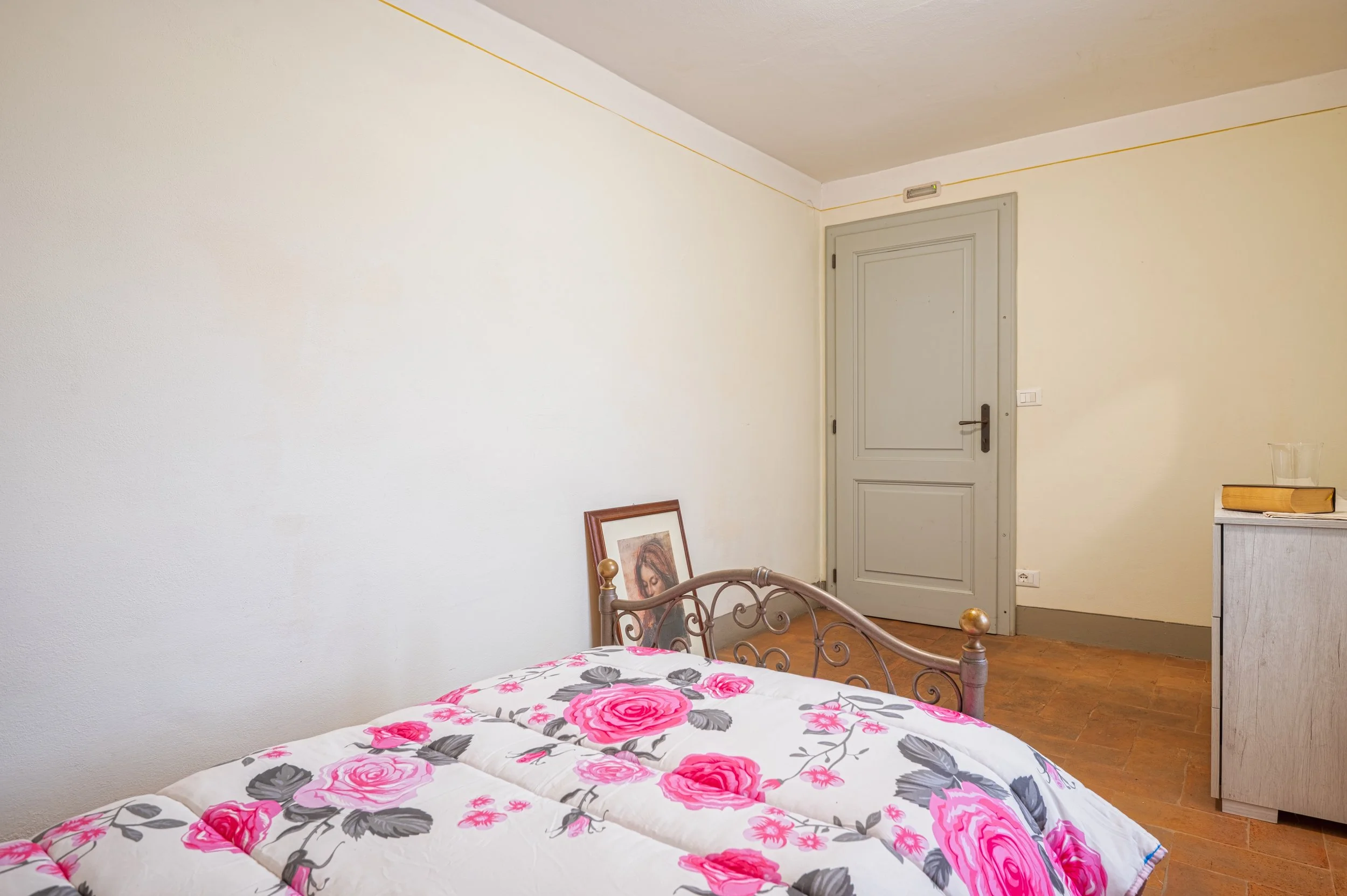 Camera da letto con letto con copriletto bianco decorato con fiori rosa e grigi, porta in legno grigia, armadio bianco, e una sedia con quadro appoggiato sopra.