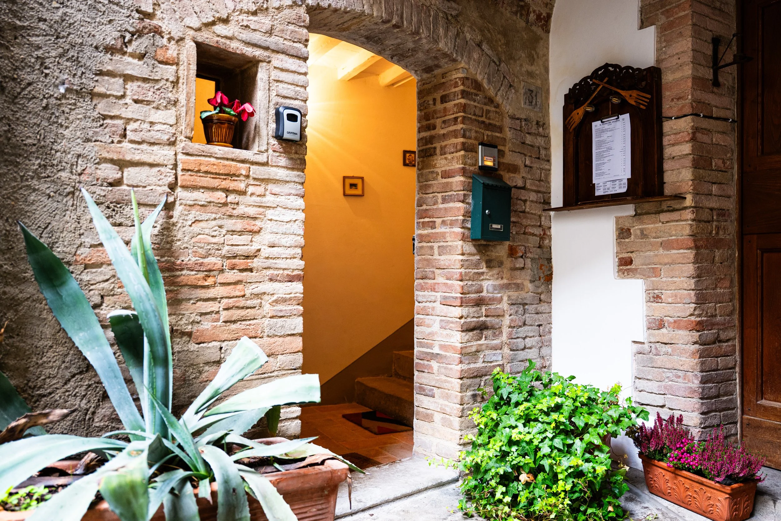 Ingresso di una casa con pareti di mattoni, piante in vaso e una scatola postale. Vano con mensola e porta con lista menu.