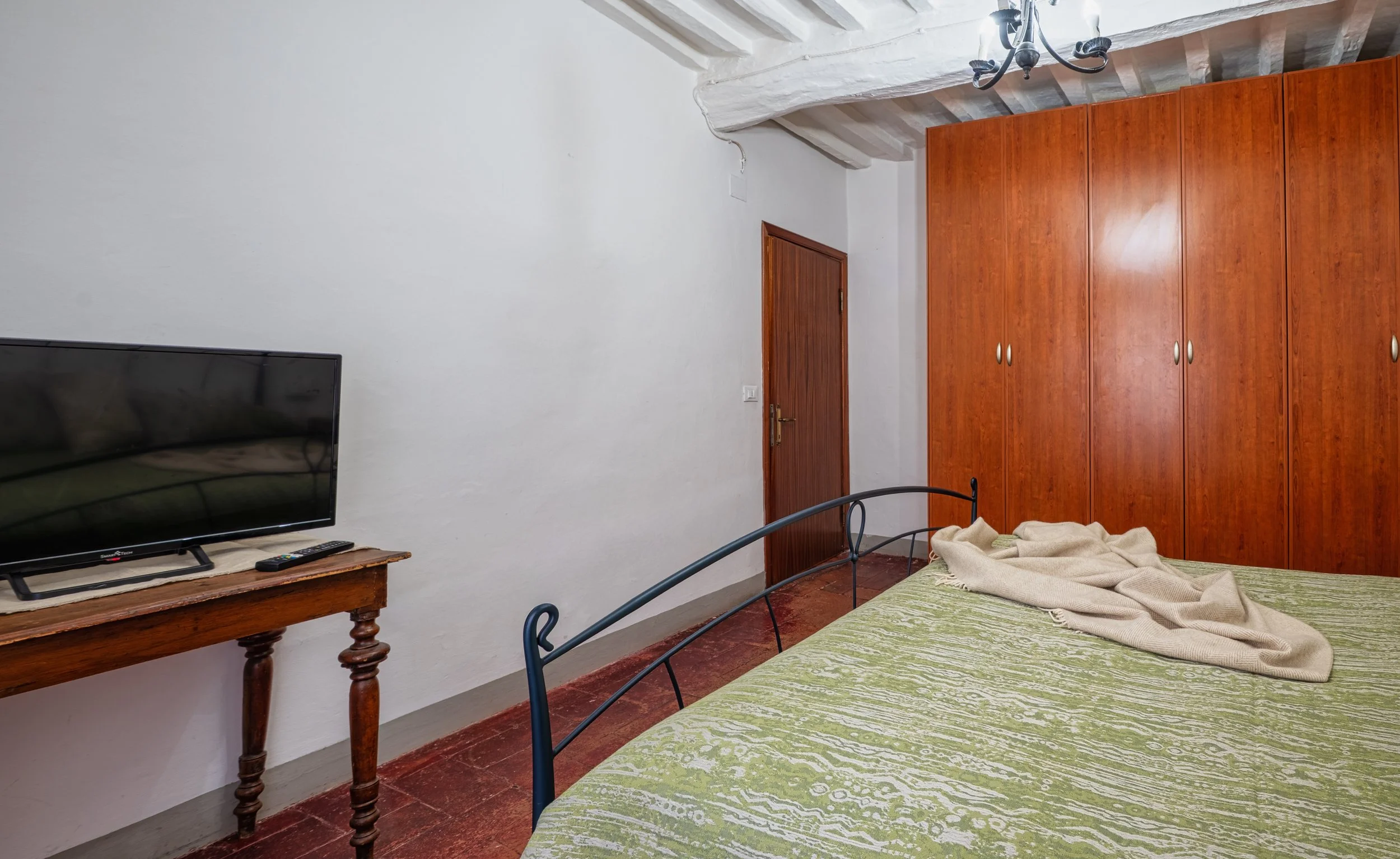 Camera con letto matrimoniale, armadio in legno, televisione su tavolo di legno, pareti bianche, porta in legno e soffitto rustico con travi di legno