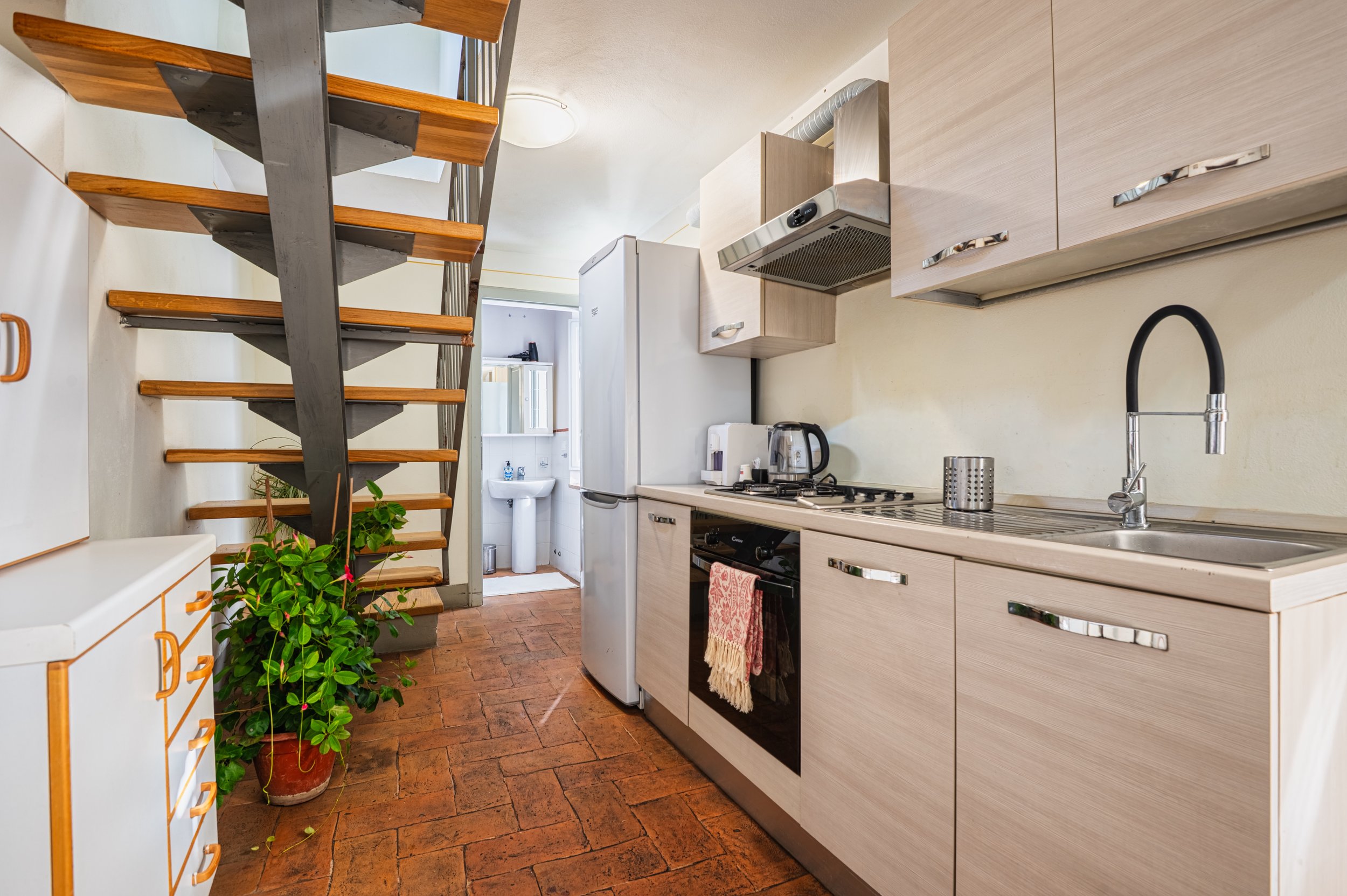 Cucina con armadi chiari, piano di lavoro, un forno, un frigorifero e un piano cottura. Scala in legno e metallo porta alla zona sopra. Pianta verde in un vaso vicino alla scala. Piccolo bagno con lavabo e specchio visibile in fondo.