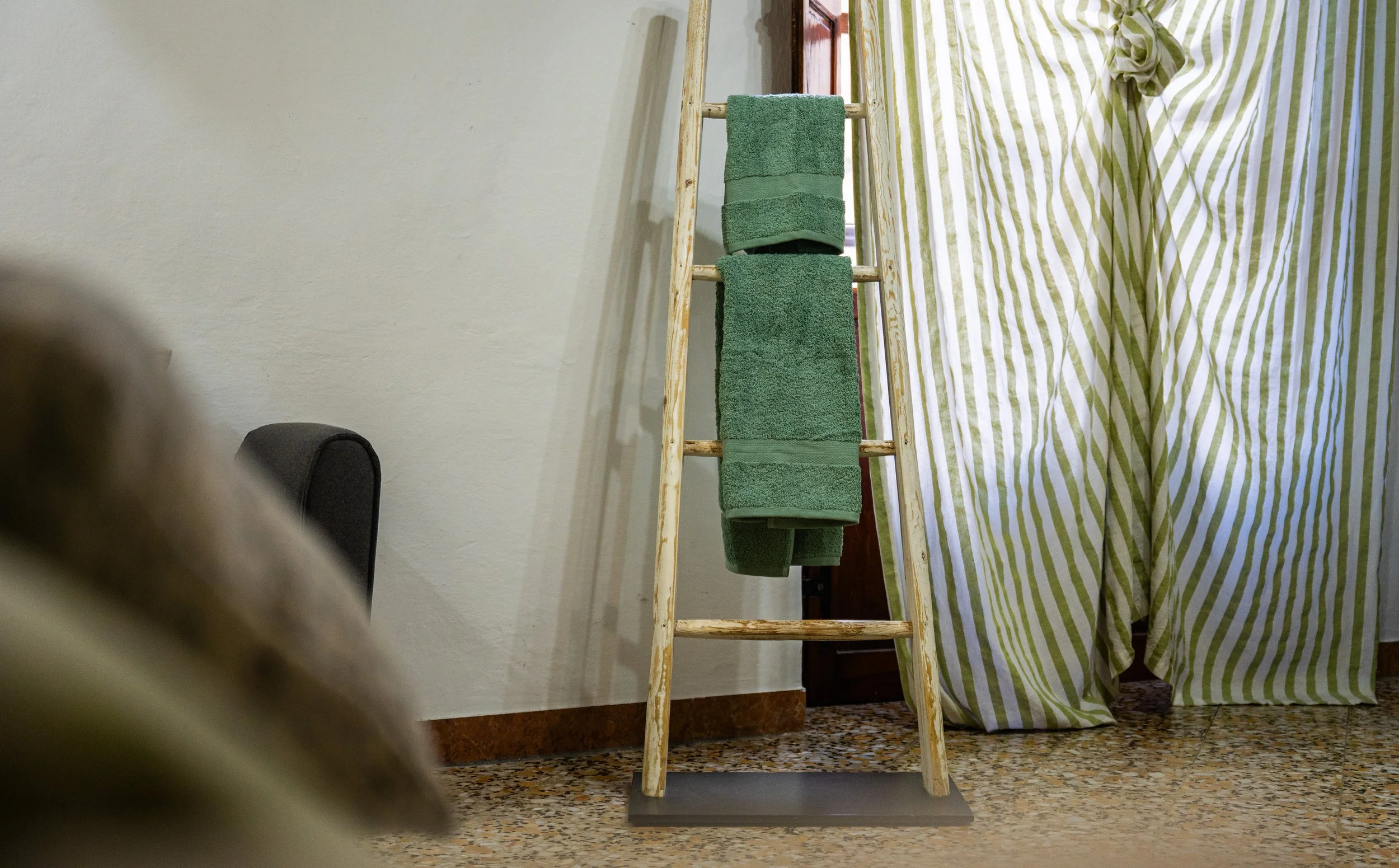 Stendino di legno con asciugamani verdi appesi, davanti a una parete bianca con tende a righe verdi e bianche.