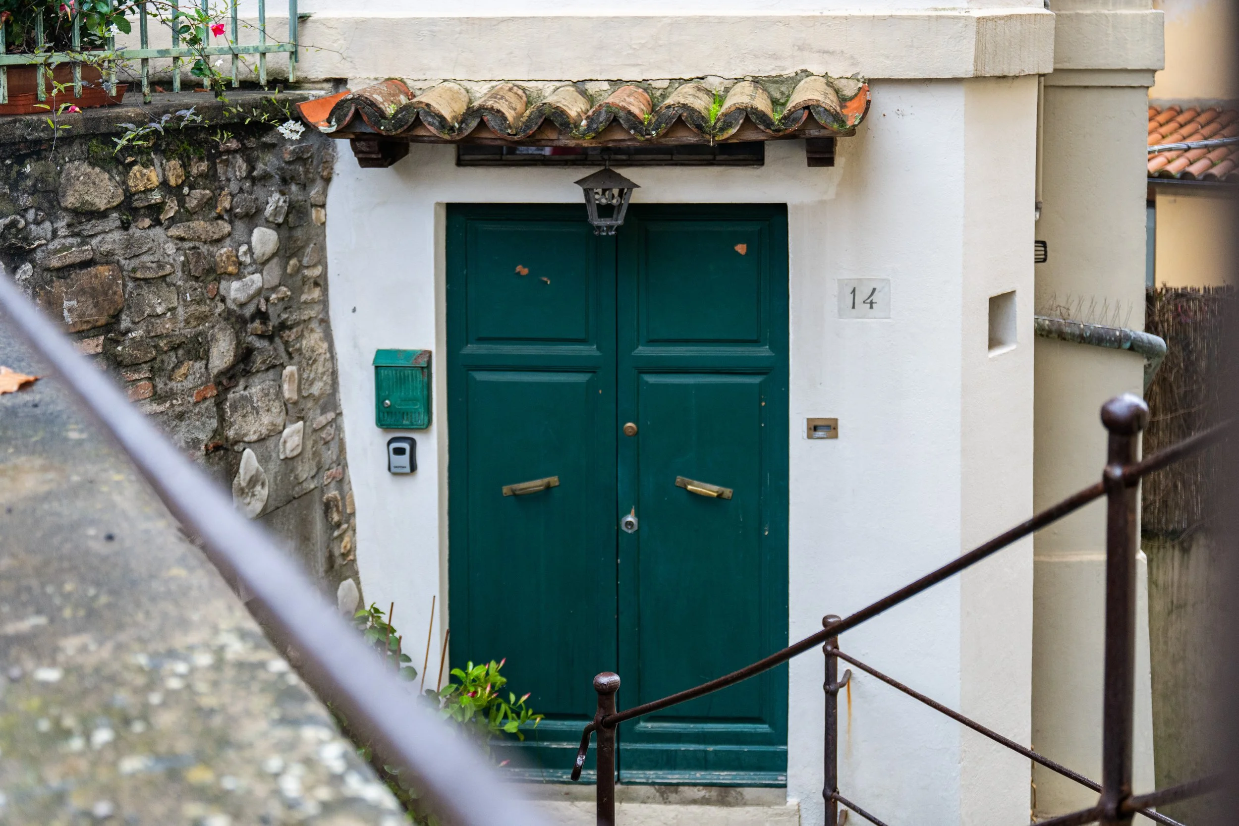 Porta verde con doppio battente, sopra un sottile tetto di tegole curve e un lampione nero appeso, vicino a una parete bianca con il numero 14.
