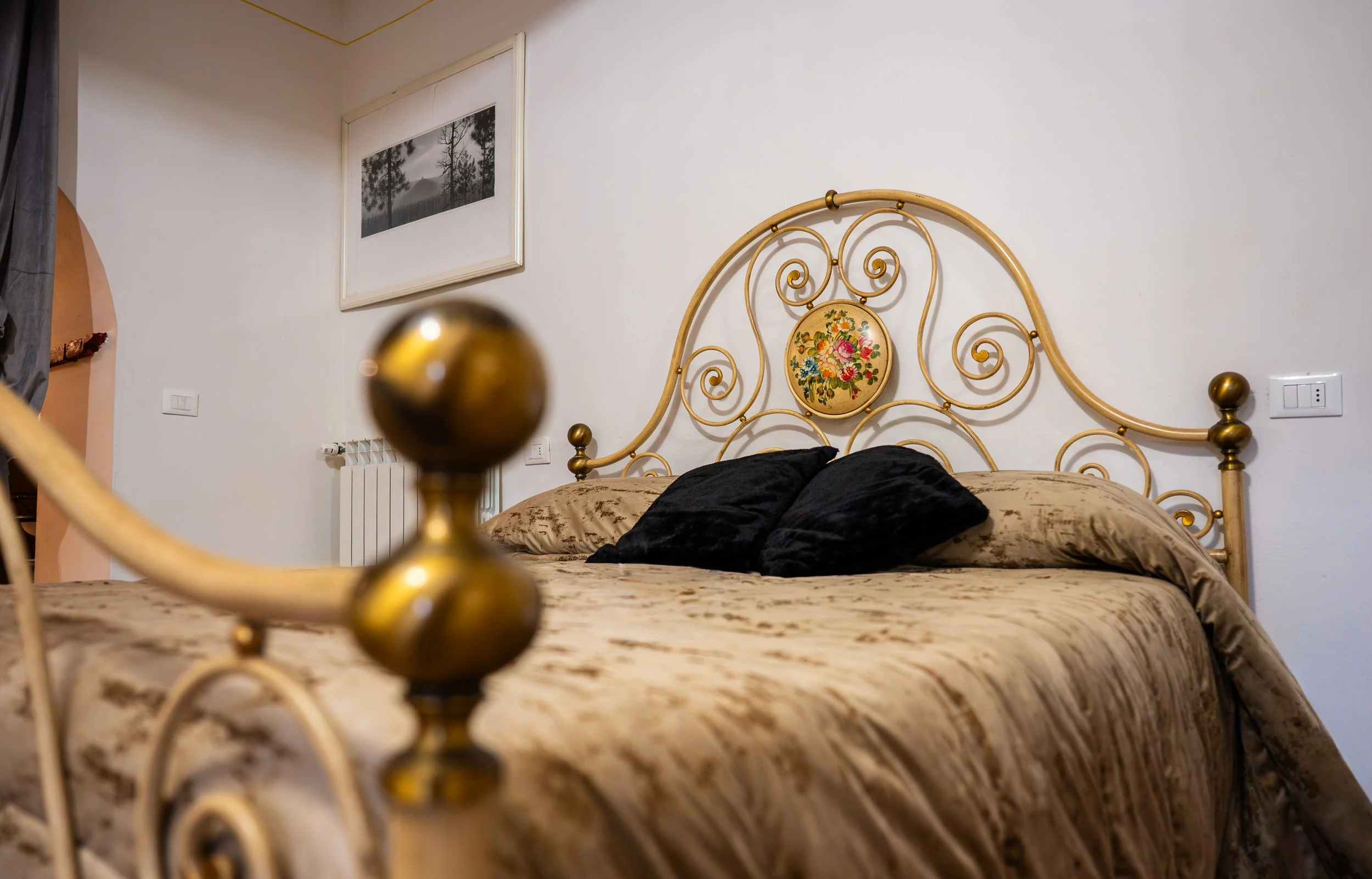 Letto matrimoniale con testiera in metallo dorato, cuscini neri, copriletto beige con motivi, quadro con paesaggio nero e bianco sopra il letto, pareti bianche, radiatore e interruttori elettrici sulla parete.